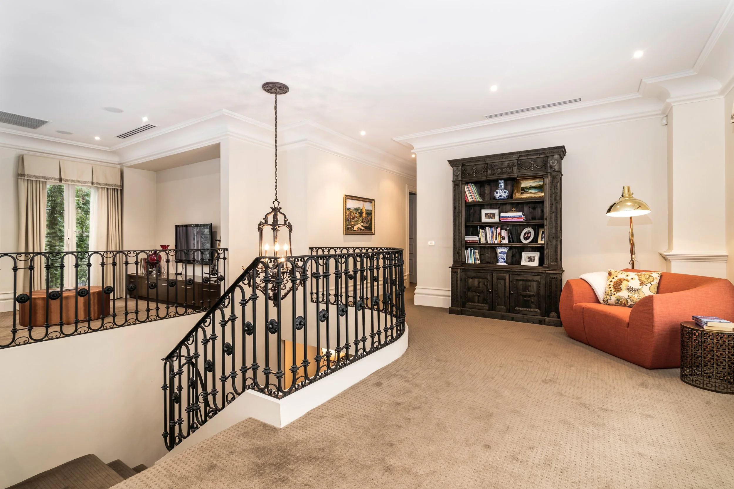 A living room with a beige carpet, white walls, and a black wrought-iron railing overlooking a staircase. There is a dark wooden bookshelf, a red sofa with a white blanket, a gold floor lamp, and a window with cream curtains.