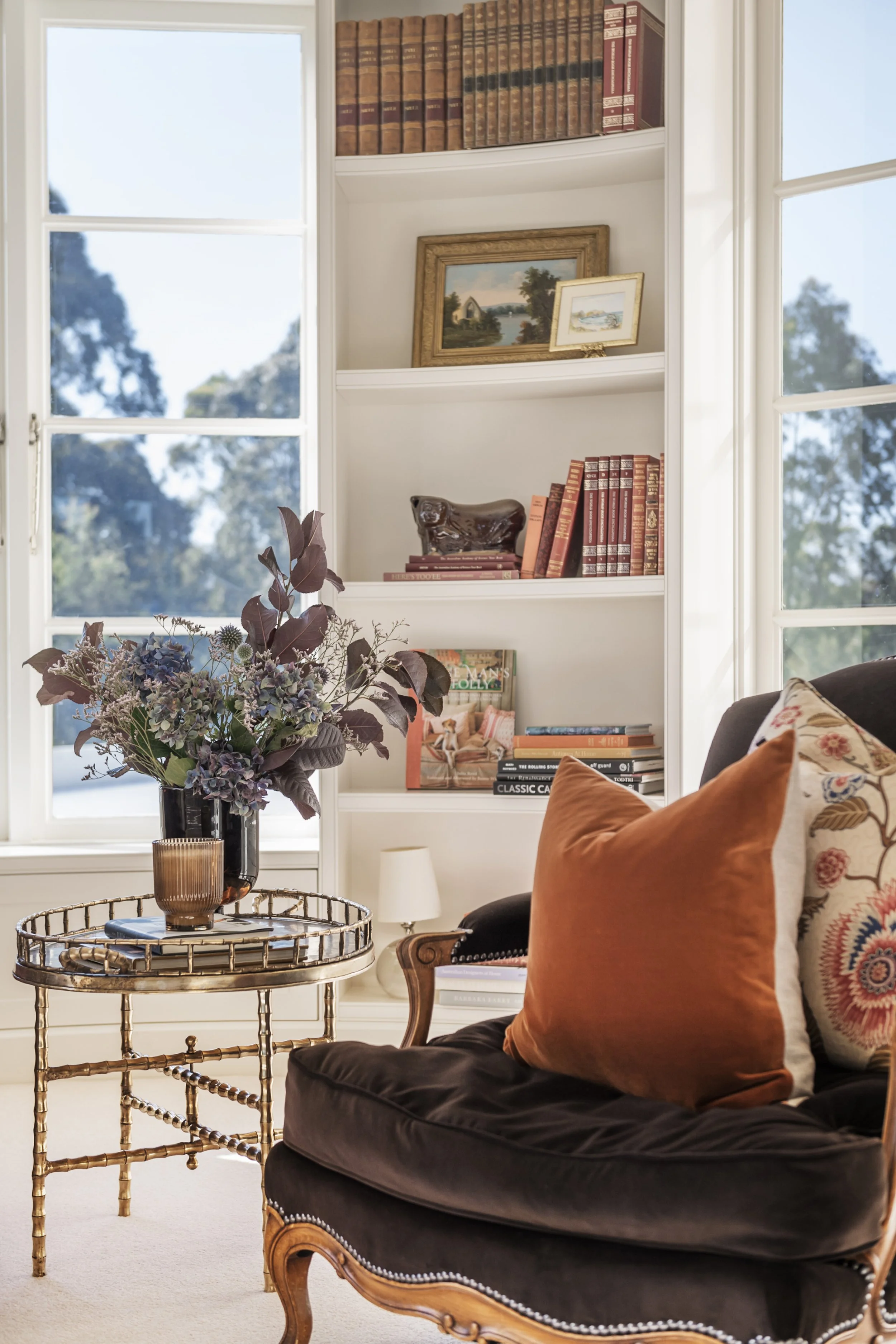 Living room with a black velvet armchair, orange pillow, gold side table with a vase of purple flowers, white bookshelf with books and framed pictures, window with a view of trees and sky.