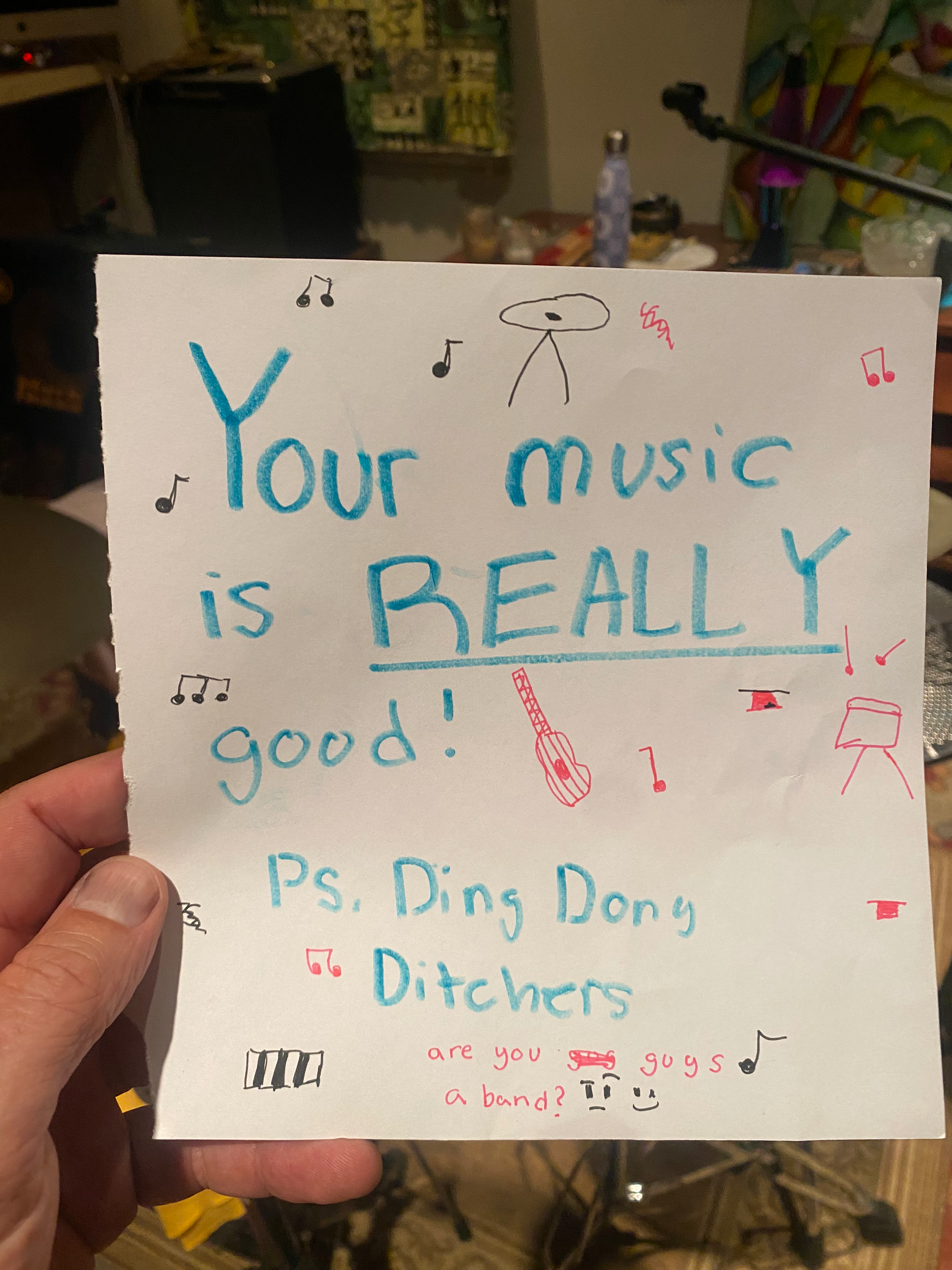 Handwritten note on white paper with musical notes, a guitar, and a small drawing of a UFO. The note reads: "Your music is really good! PS. Ding dong DITCHERS are you guys a band? 😊" in large blue and red letters.