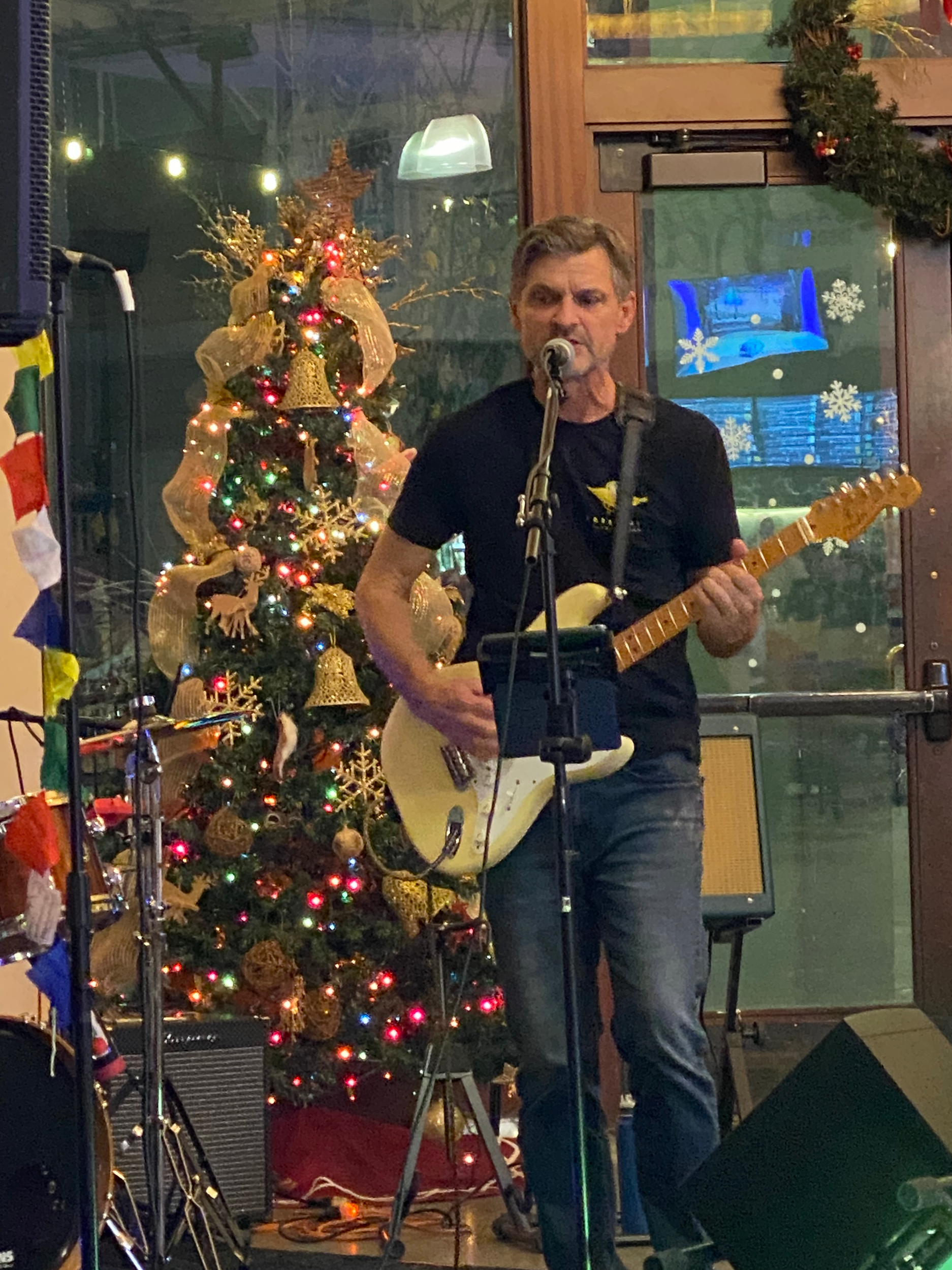 Johnny Slide from 8 track mind playing an electric guitar and singing into a microphone on stage in San Jose, Ca. , with a decorated Christmas tree in the background.