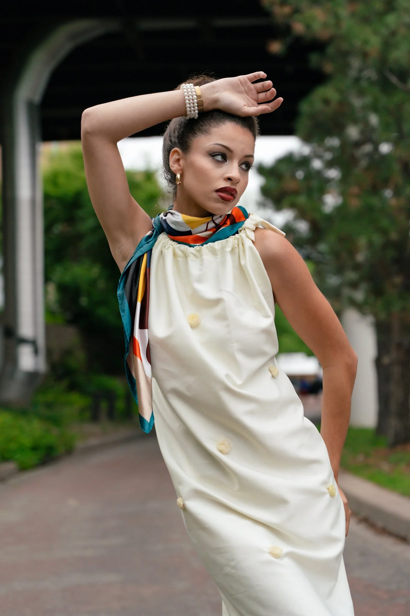 A woman wearing a cream dress with decorative buttons, a colorful scarf around her neck, and a pearl bracelet, striking a pose outdoors near a bridge with greenery in the background.