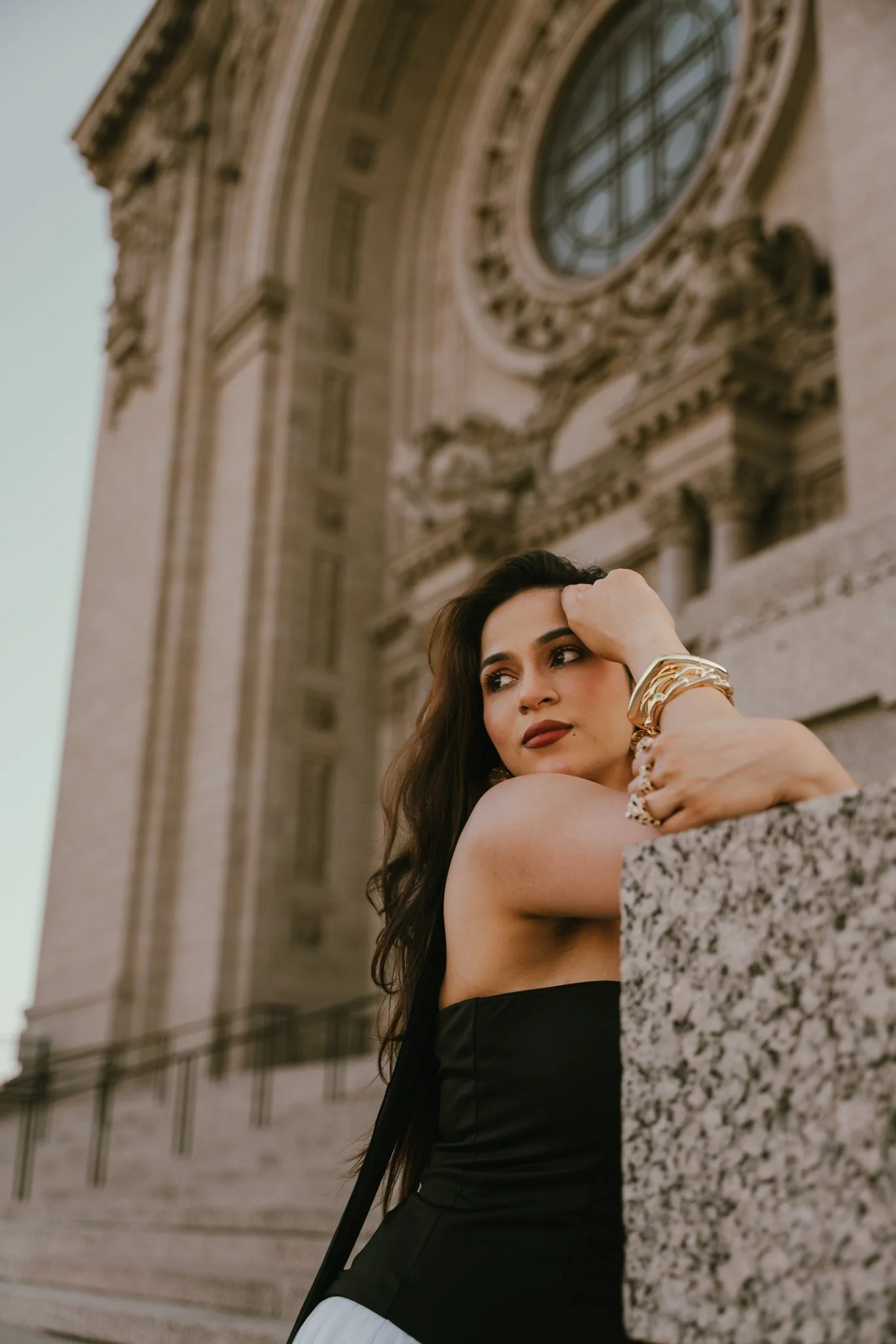 A woman with long dark hair and wearing gold jewelry relaxes against a stone railing outside a historic building with intricate architectural details and a large clock above.