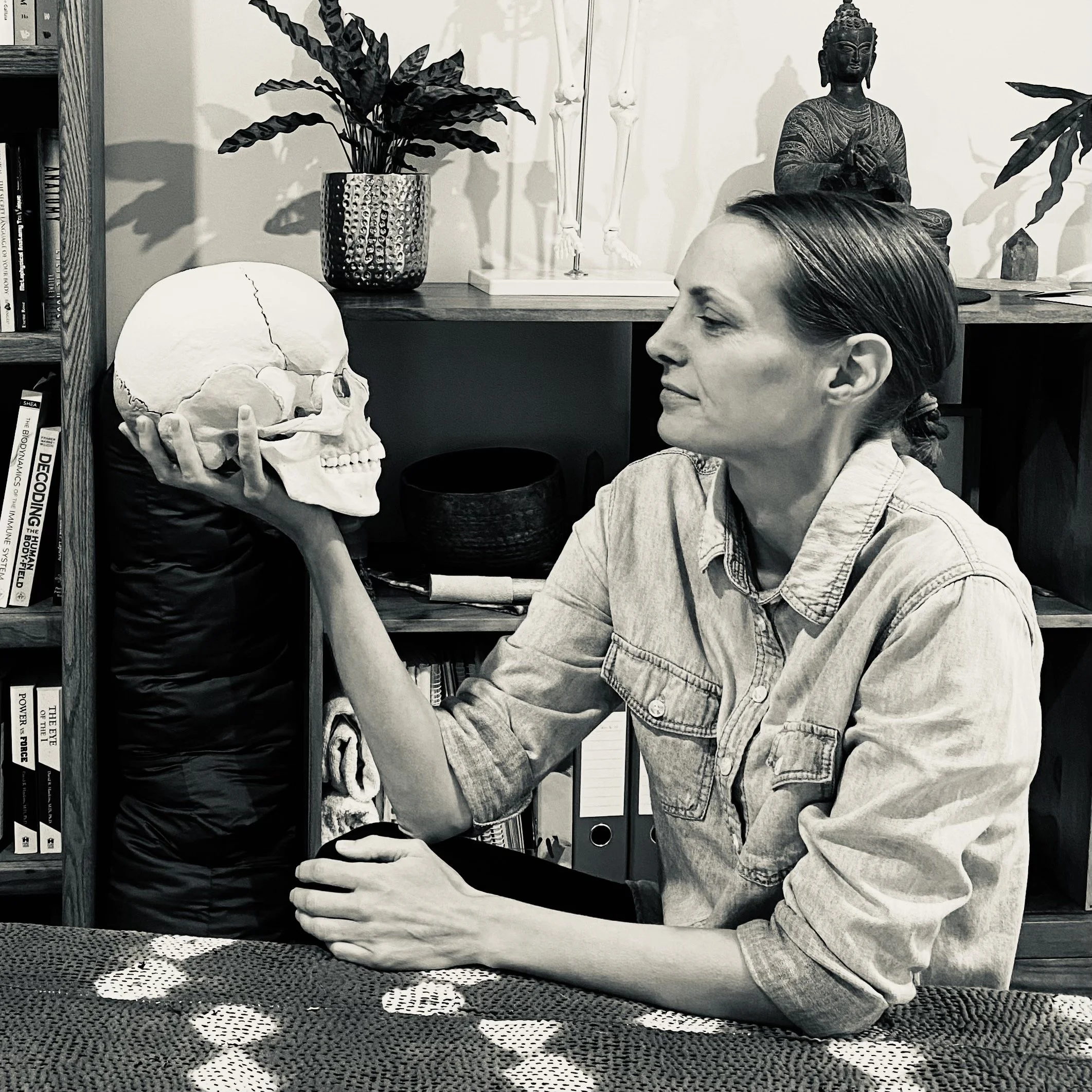 A woman sitting on the floor in front of a skull, holding its chin with her hand. The scene includes books on a shelf, a plant in a metallic pot, a bass skeleton model, and a Buddha statue in the background, all set within a room.