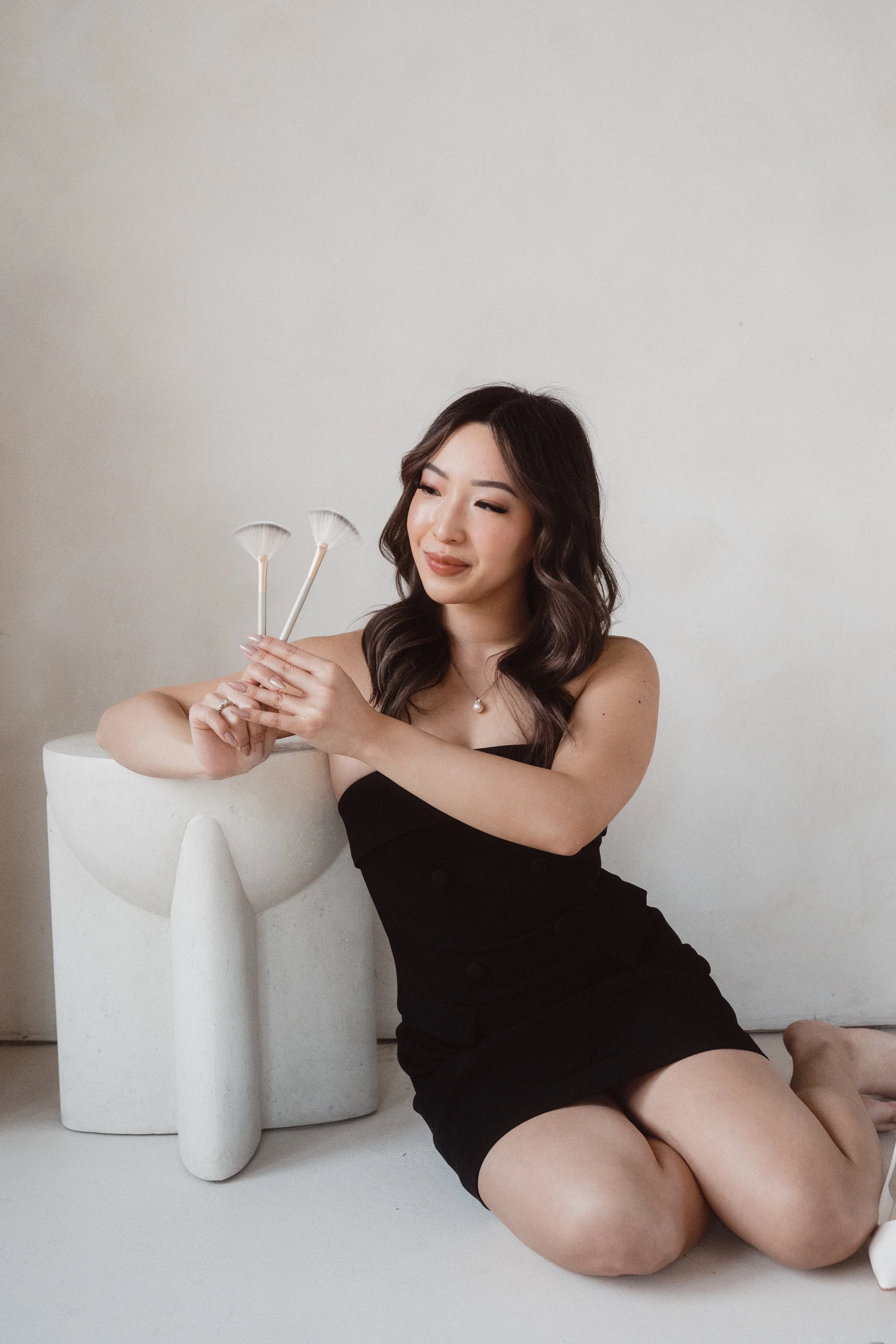 Woman with wavy dark hair in a black strapless dress sitting on the floor, holding three small white brushes next to a modern white sculpture, against a plain light background.