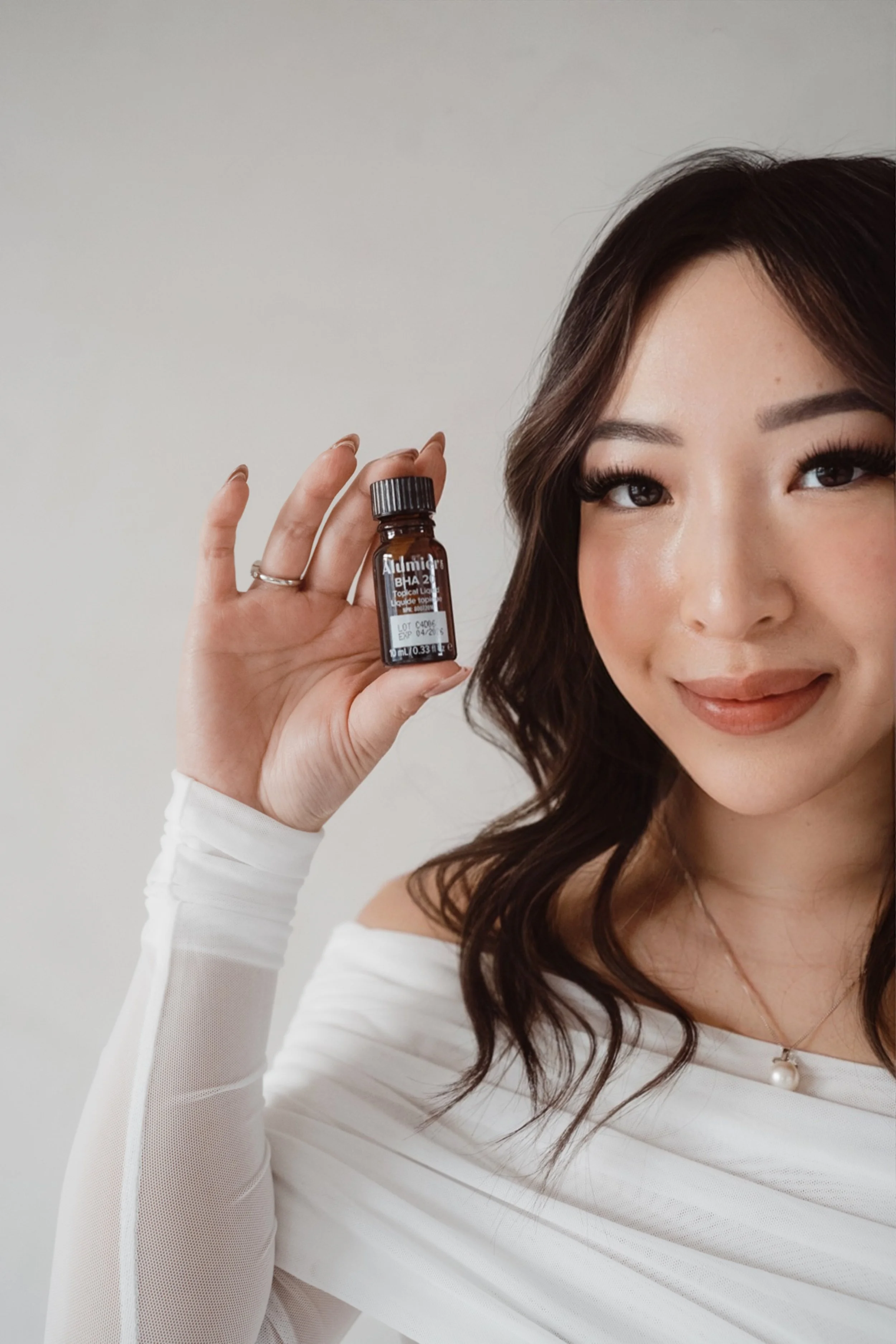 A woman with dark brown hair holding a small brown bottle labeled 'Admidar BHA 2%'. She is smiling and wearing an off-shoulder white top and a pearl necklace.