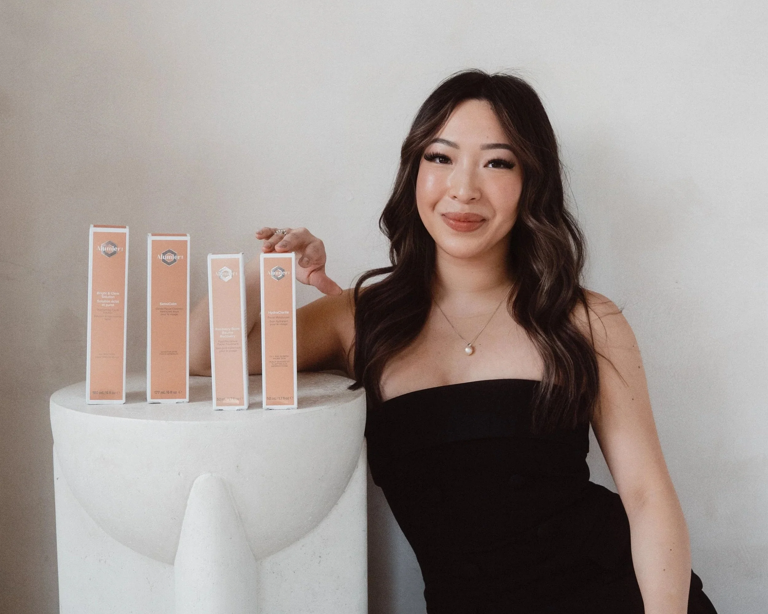 A woman in a black strapless top with wavy brown hair and a pearl necklace standing next to a white pedestal with four skincare product boxes on top.