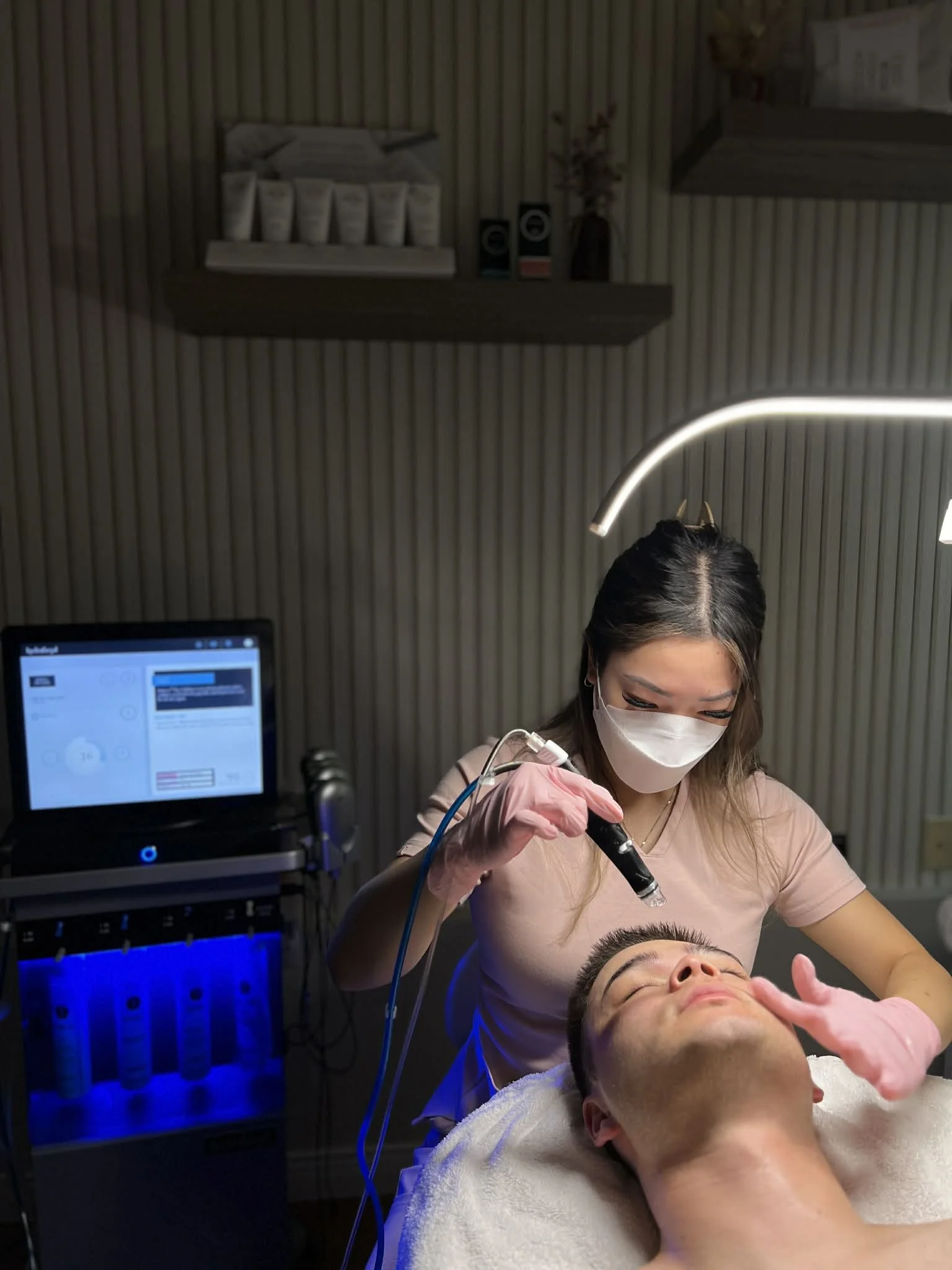 A woman wearing a mask and gloves is performing a facial treatment on a man lying on a treatment bed with his eyes closed in a skincare clinic.