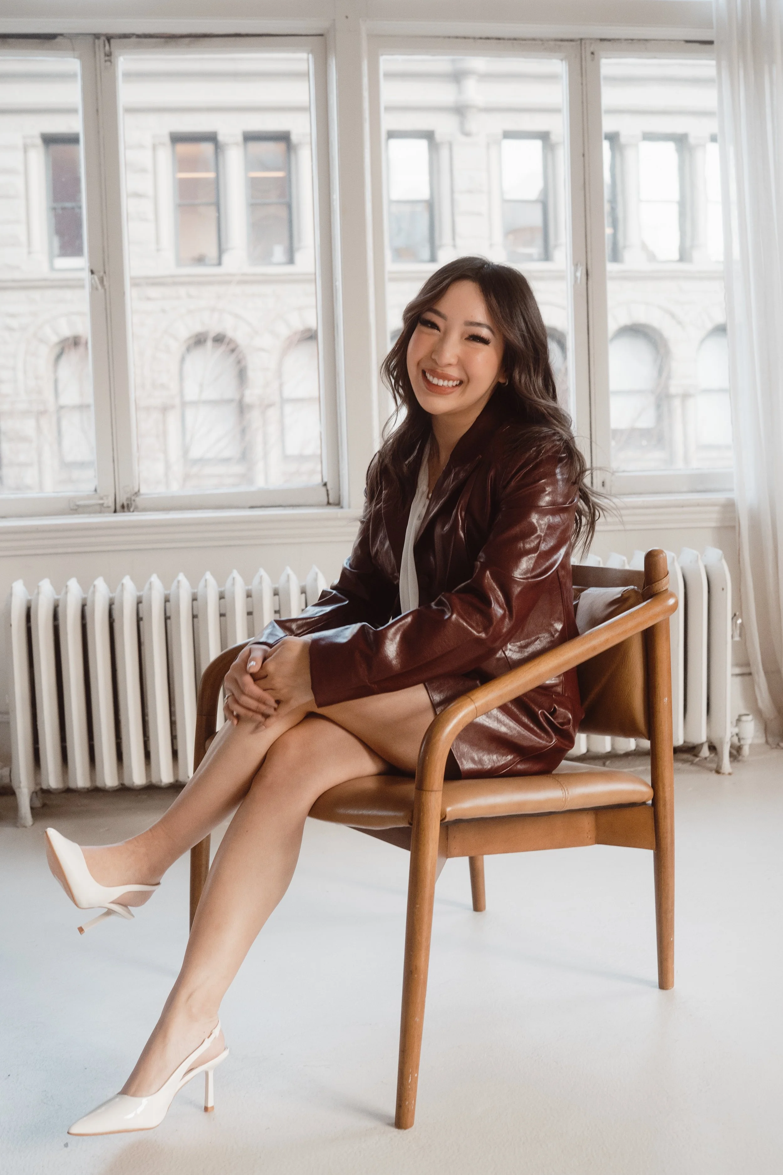 A woman sitting on a wooden chair in front of large windows with an urban cityscape view, smiling and wearing a brown leather jacket, white dress, and white high heels.