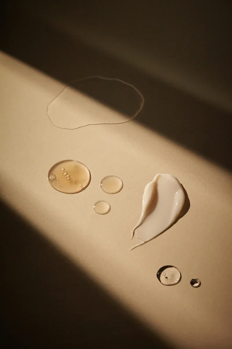 Close-up of water droplets and a dollop of cream or lotion on a beige surface, with shadowed lighting.