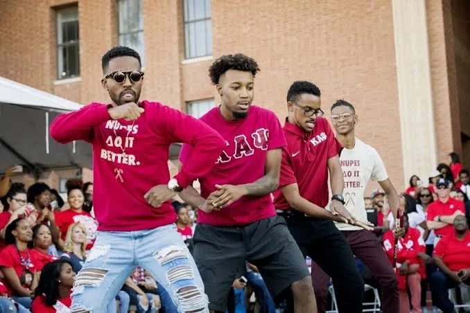 Four young men dancing outside at a college event, with a crowd of people sitting and standing behind them.