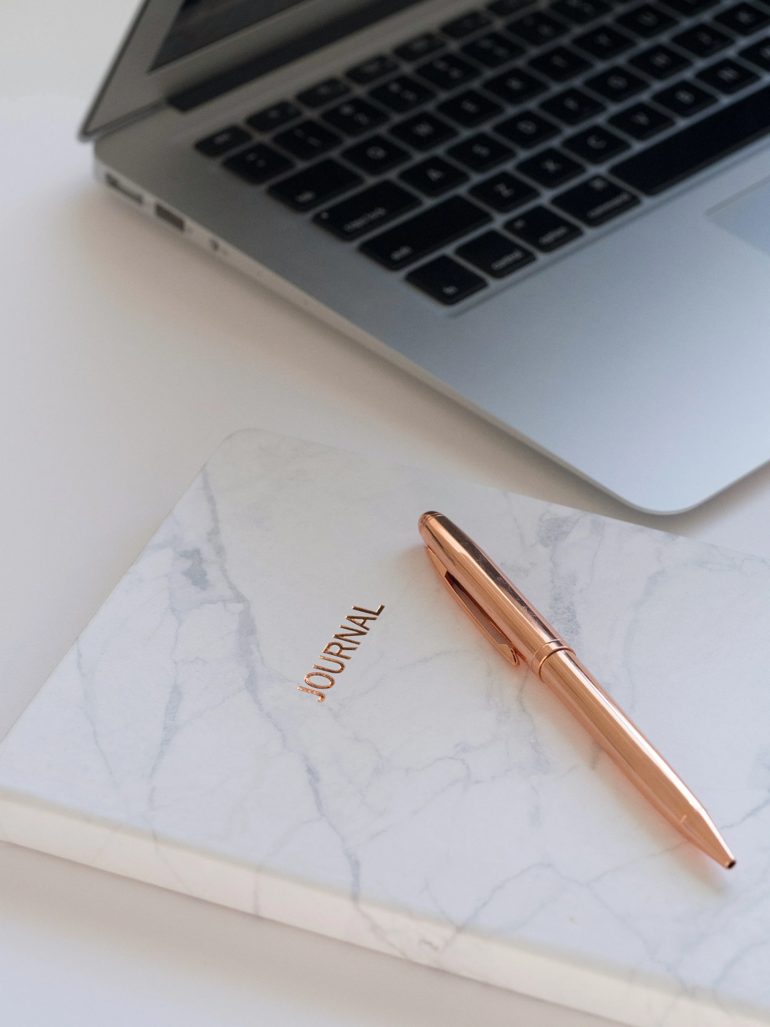 A marble-covered journal with the word 'JOURNAL' etched in gold, a rose gold pen resting on it, and a laptop nearby on a white surface.
