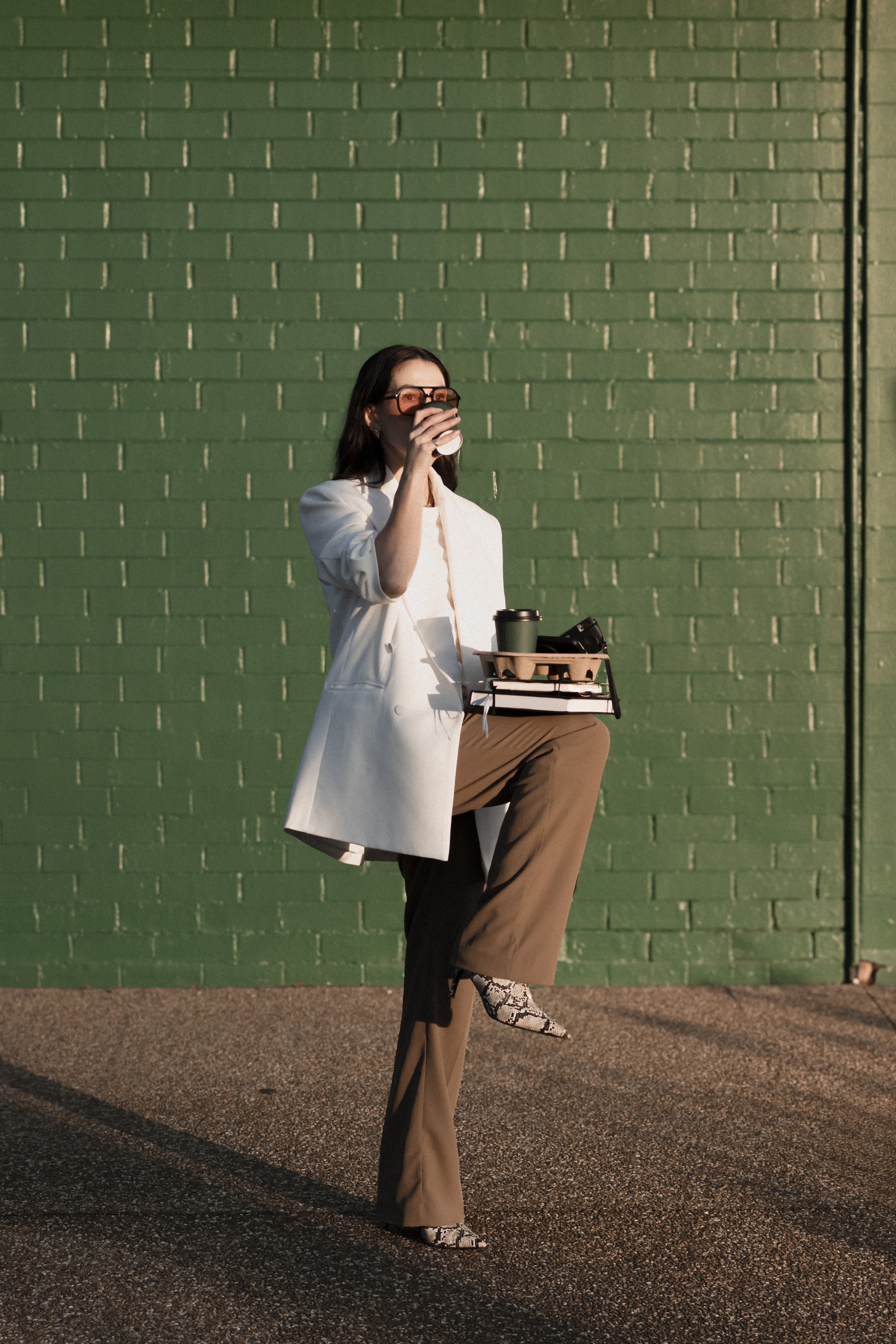 A woman with dark hair wearing sunglasses, a white blazer, and beige pants stands against a green brick wall, holding a coffee cup and a tray with a drink, a camera, and a notebook, with a snake print shoe.