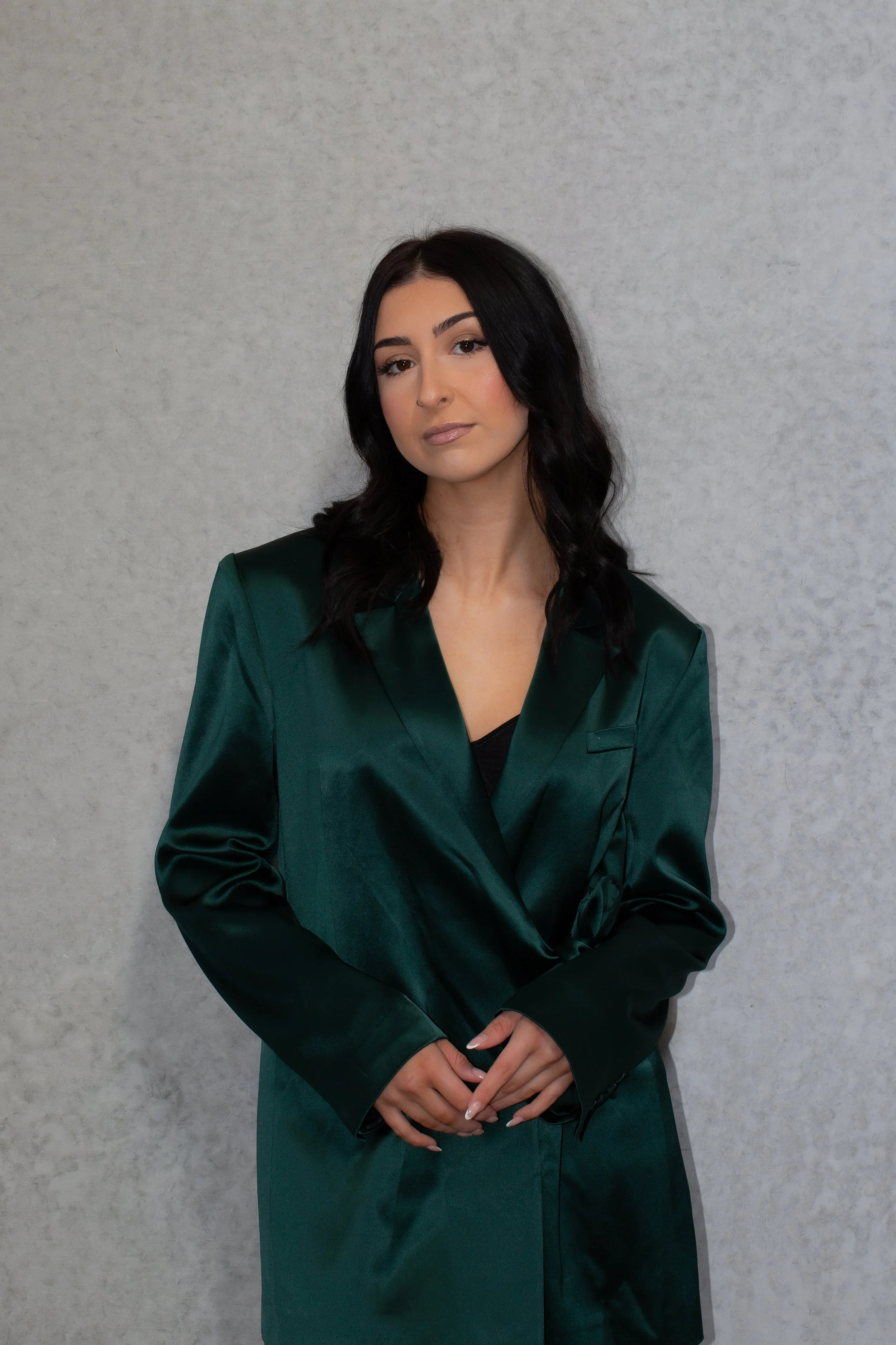 A young woman with long black hair standing against a plain light gray background, wearing a dark green satin blazer.