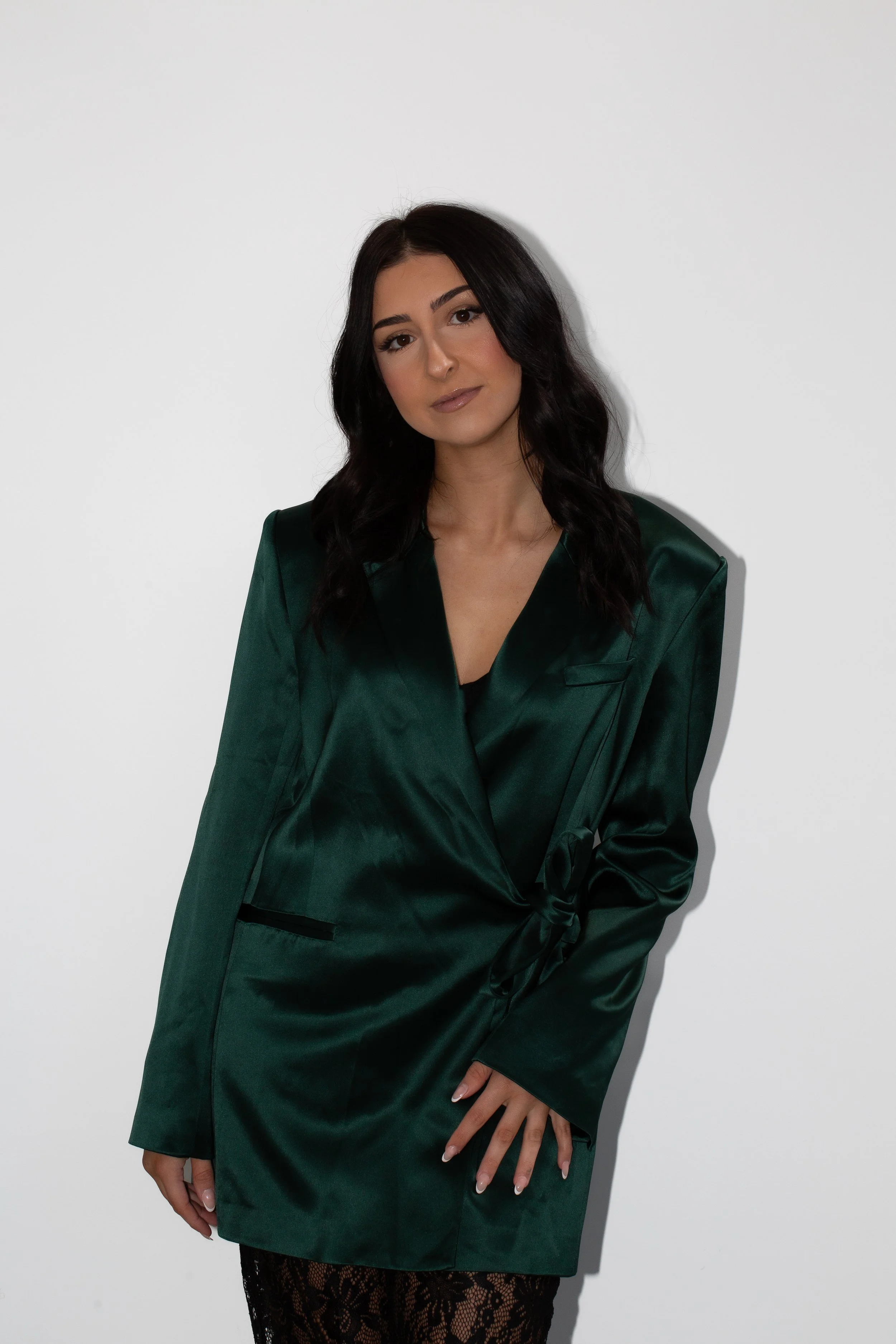 A young woman with long dark hair, wearing a emerald green satin blazer and a matching skirt with lace trim, standing against a plain white wall.