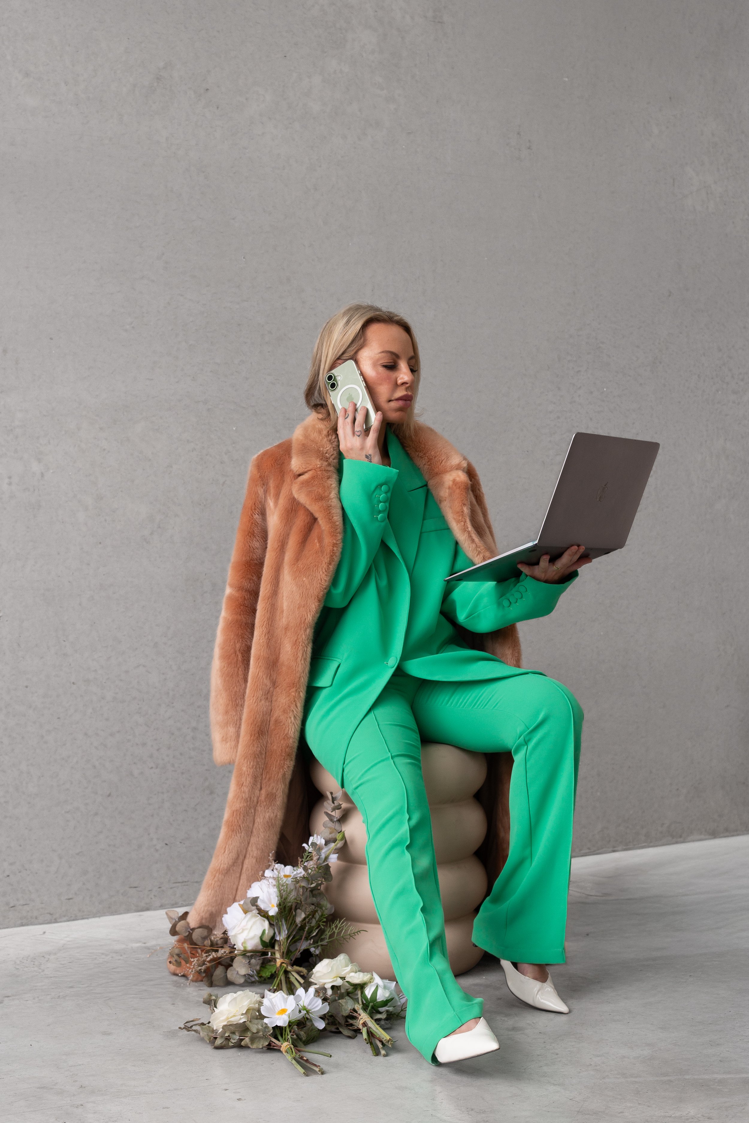 A woman in a green suit and white heels sitting on a small stool, holding a laptop and talking on a phone, with flowers on the floor and a fur coat draped over her shoulders.