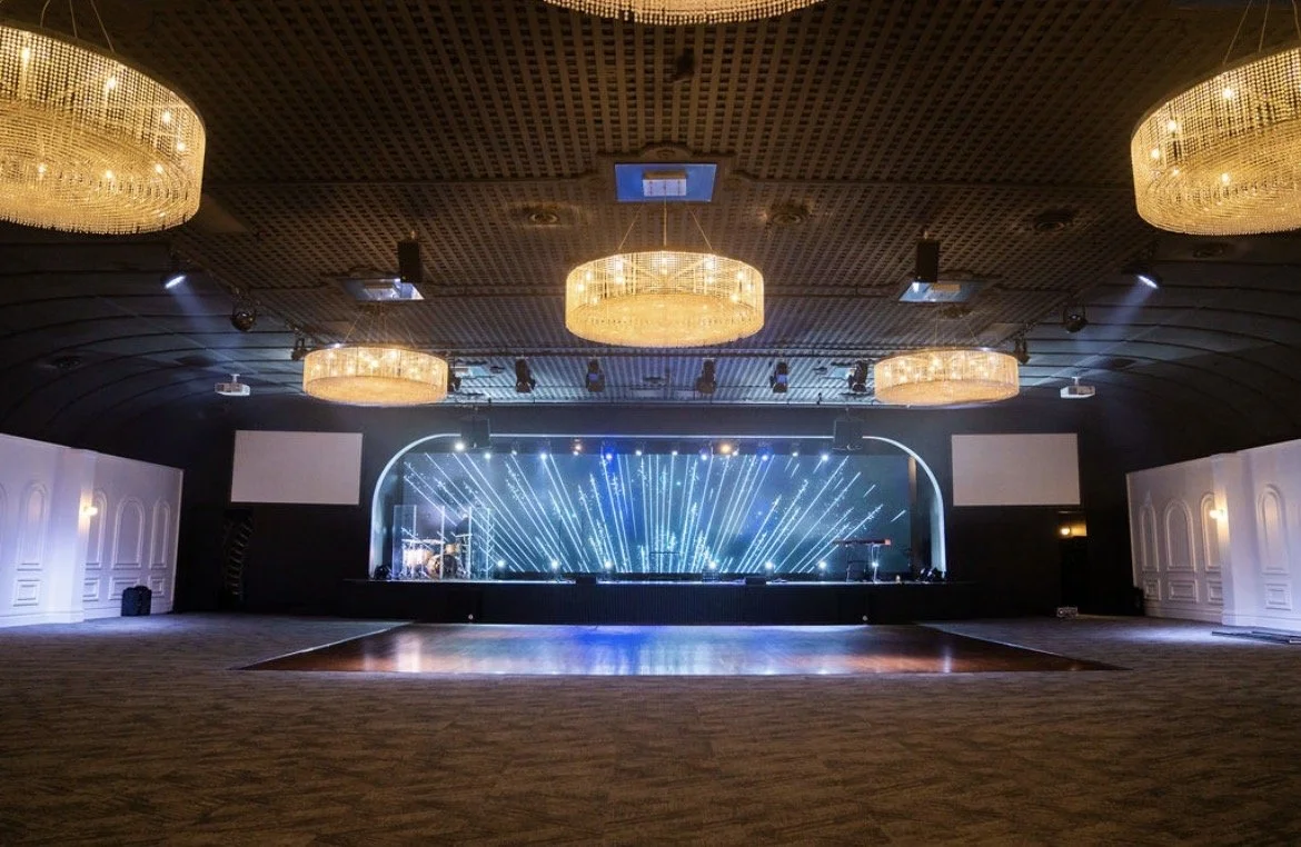 Empty event hall with a stage, illuminated by blue and white laser light effects, and five large ornate chandeliers hanging from a wooden ceiling.