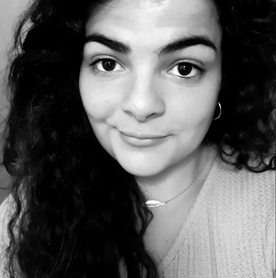 A young woman with curly dark hair, light skin, and wearing hoop earrings, a necklace with a pendant, and a textured top, smiling at the camera in a black and white photo.