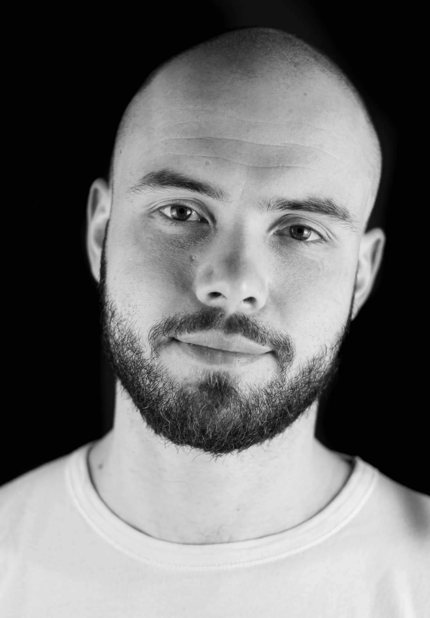 Black and white portrait of a young man with a beard, short hair, and light-colored eyes, wearing a light T-shirt, against a dark background.