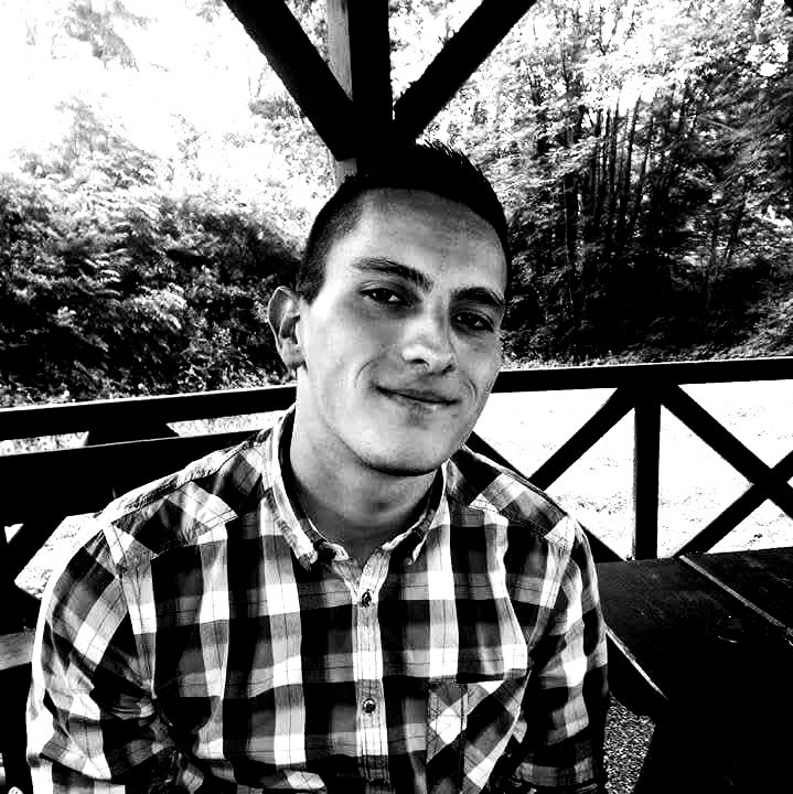 A young man wearing a checkered shirt sitting outdoors on a wooden deck with railing, with trees and foliage in the background.