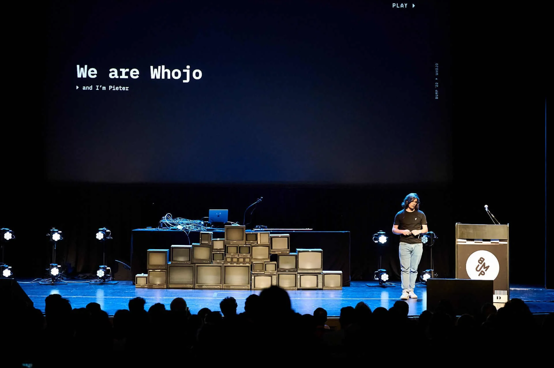 A man stands on stage next to a podium with a microphone. Behind him, a large screen displays the text 'We are Whojo and I'm Pieter.' The stage features a structure made of stacked vintage televisions and monitors. Stage lights illuminate the scene, and audience members are visible as silhouettes at the front.