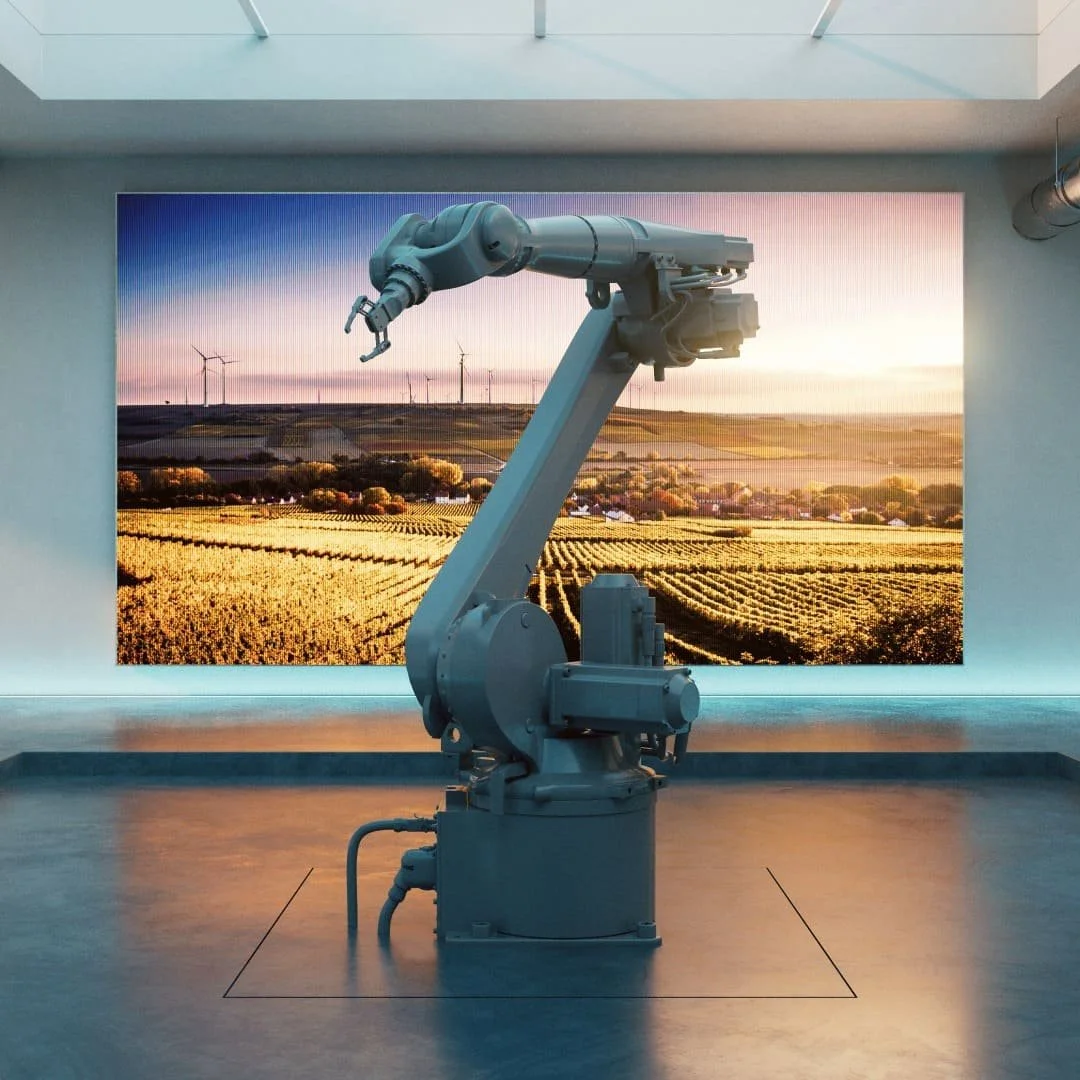 Industrial robotic arm in front of a large digital screen displaying a landscape with wind turbines and farmland at sunset.