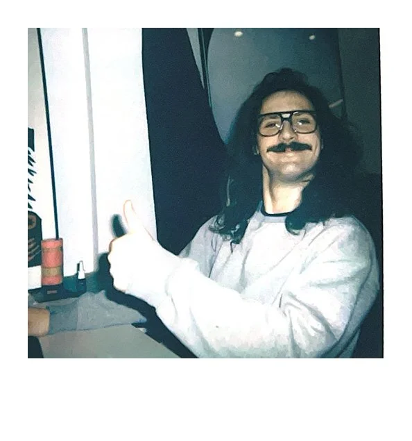 Person wearing glasses and a fake mustache, giving a thumbs-up while sitting at a desk with art supplies like glue and paint, in what appears to be a creative workspace.