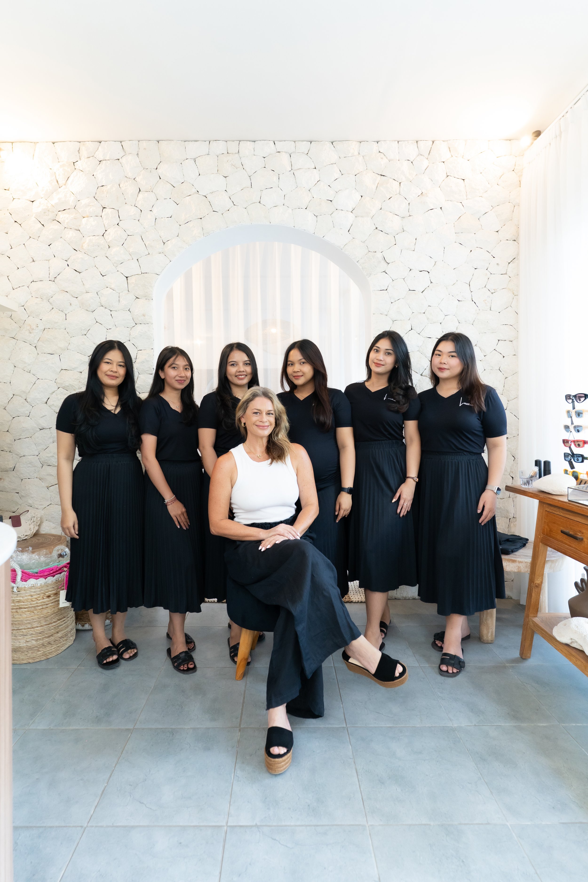 Group of seven women, one seated in front and six standing behind, posing for a photo indoors with a white stone wall background.