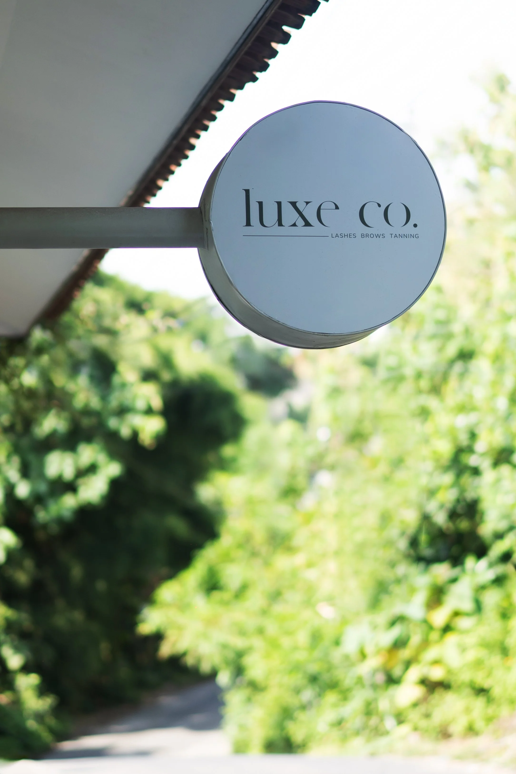 Sign for Luxe Co. business, offering eyelashes, eyebrows, and tanning services, showing the sign attached to the building's exterior with a cloudy sky and trees in the background.
