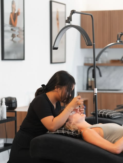 A woman receiving a beauty treatment in a salon, lying on a black bed with a pillow, with a technician applying a procedure near her face.