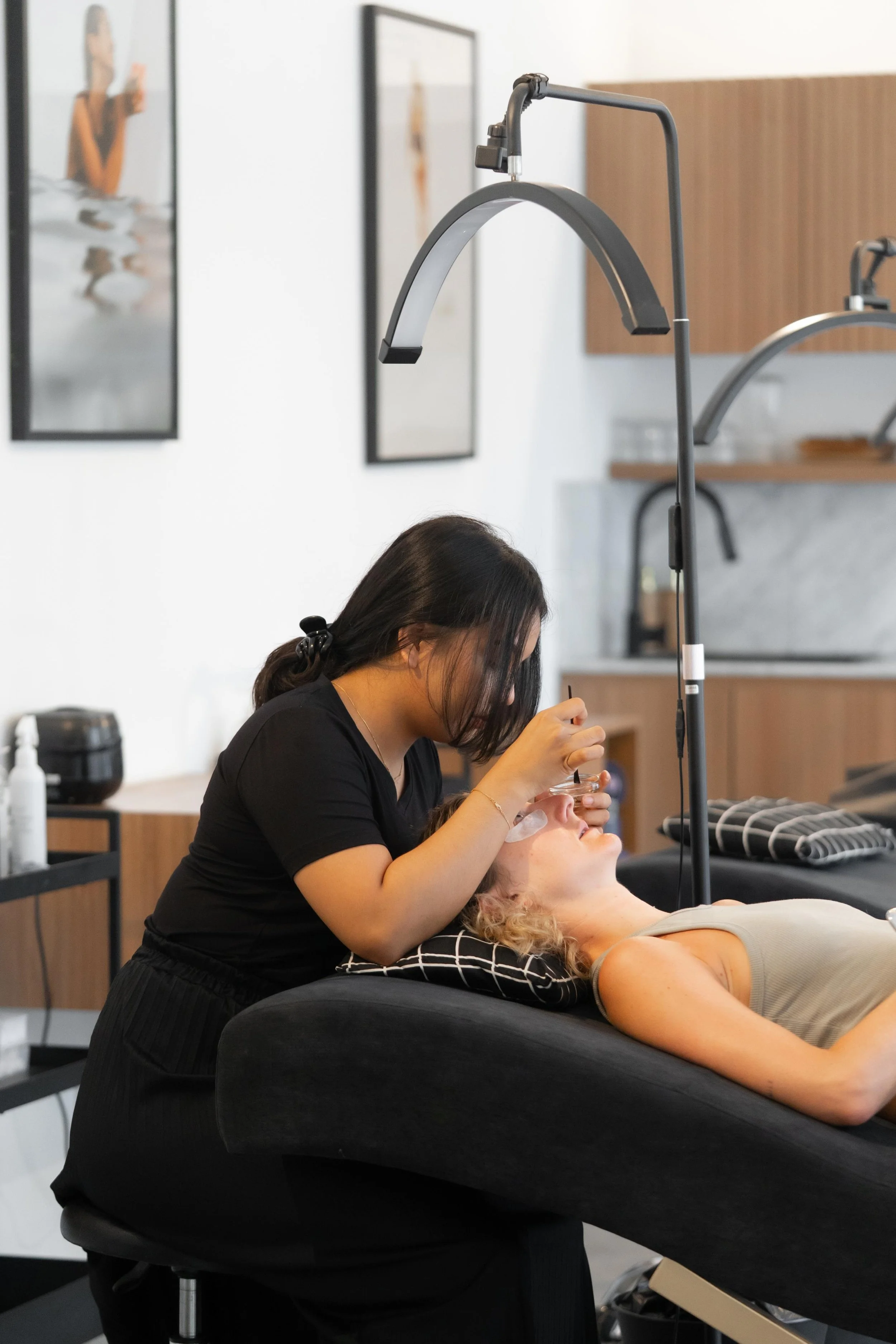A woman receiving a beauty treatment at a salon, lying on a black treatment bed while a technician applies a product near her eyes.