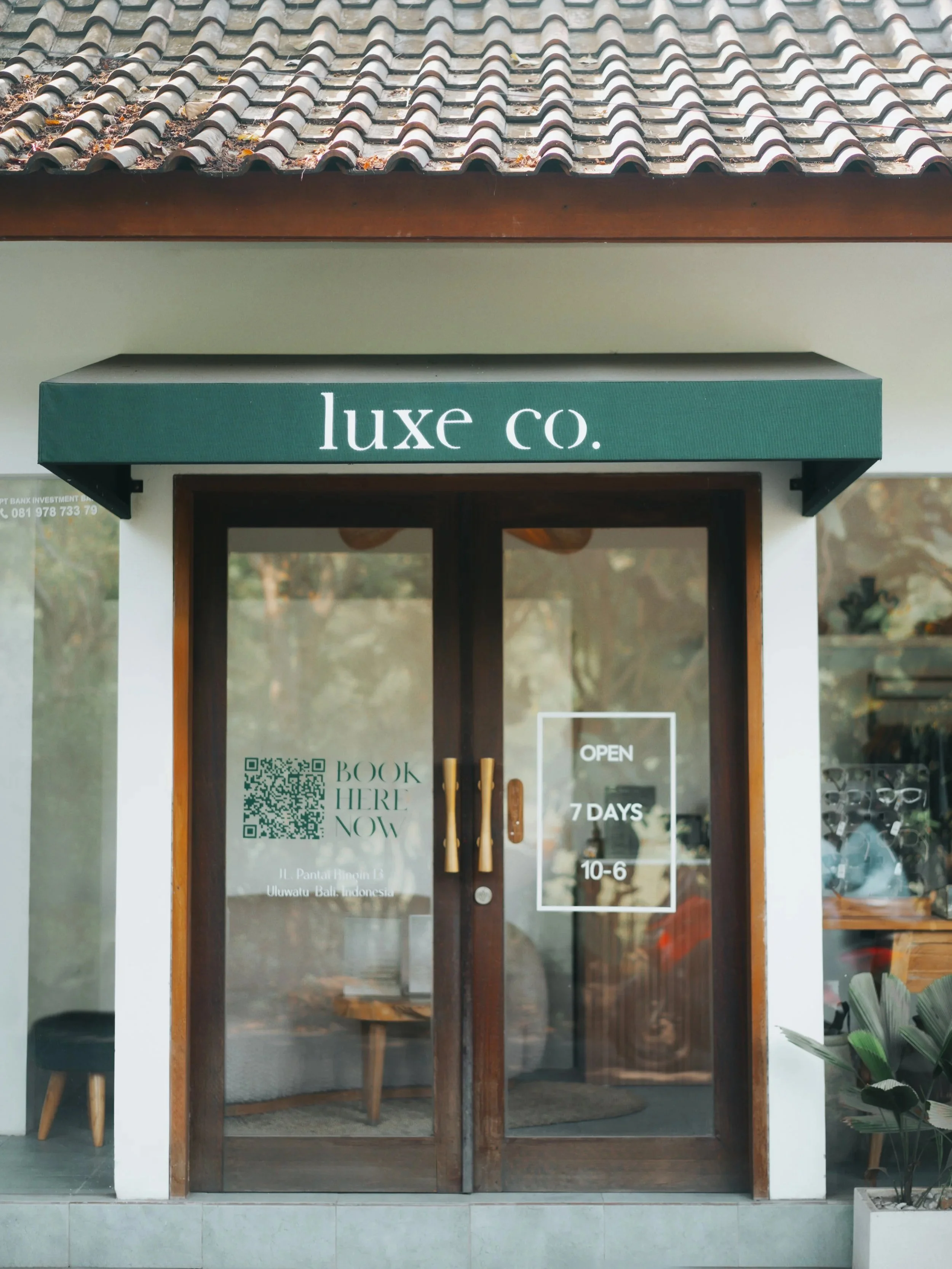 The entrance of a boutique shop with a green awning labeled 'luxe co.' above glass doors. The shop offers booking, is open 7 days, from 10 am to 6 pm, and displays a QR code and address on the door.