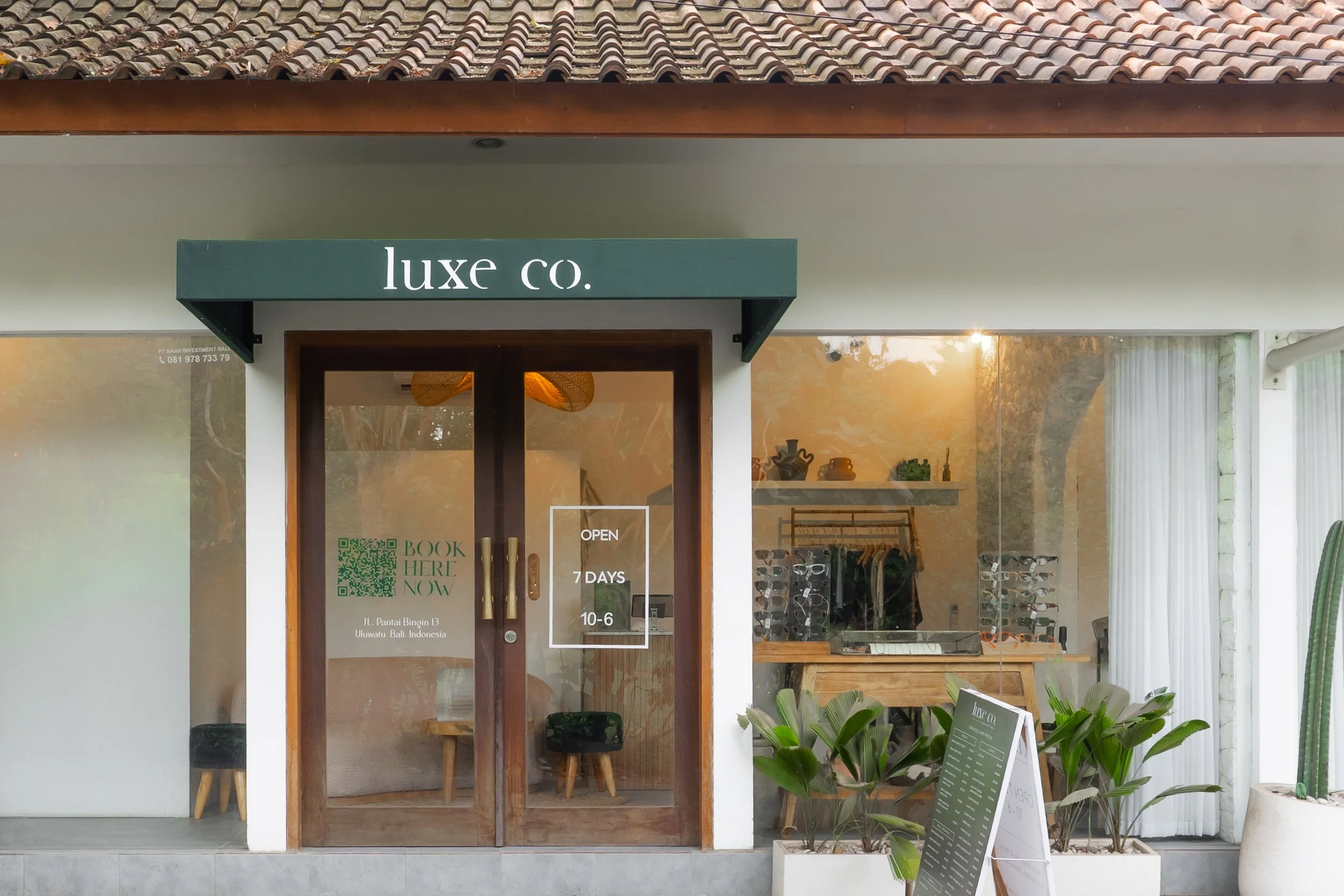 The storefront of a shop named Luxe Co. with a glass door and windows, displaying the shop's hours of operation and a QR code. Inside, there are shelves with plants and decorative items, and a clothing rack is visible. Potted plants are outside the shop.
