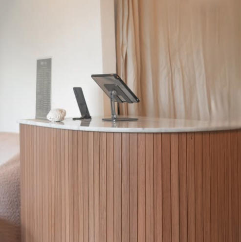 A curved wooden counter with a small stand holding a tablet, a black phone, a textured decorative object, and a tall speaker in a room with beige curtains.