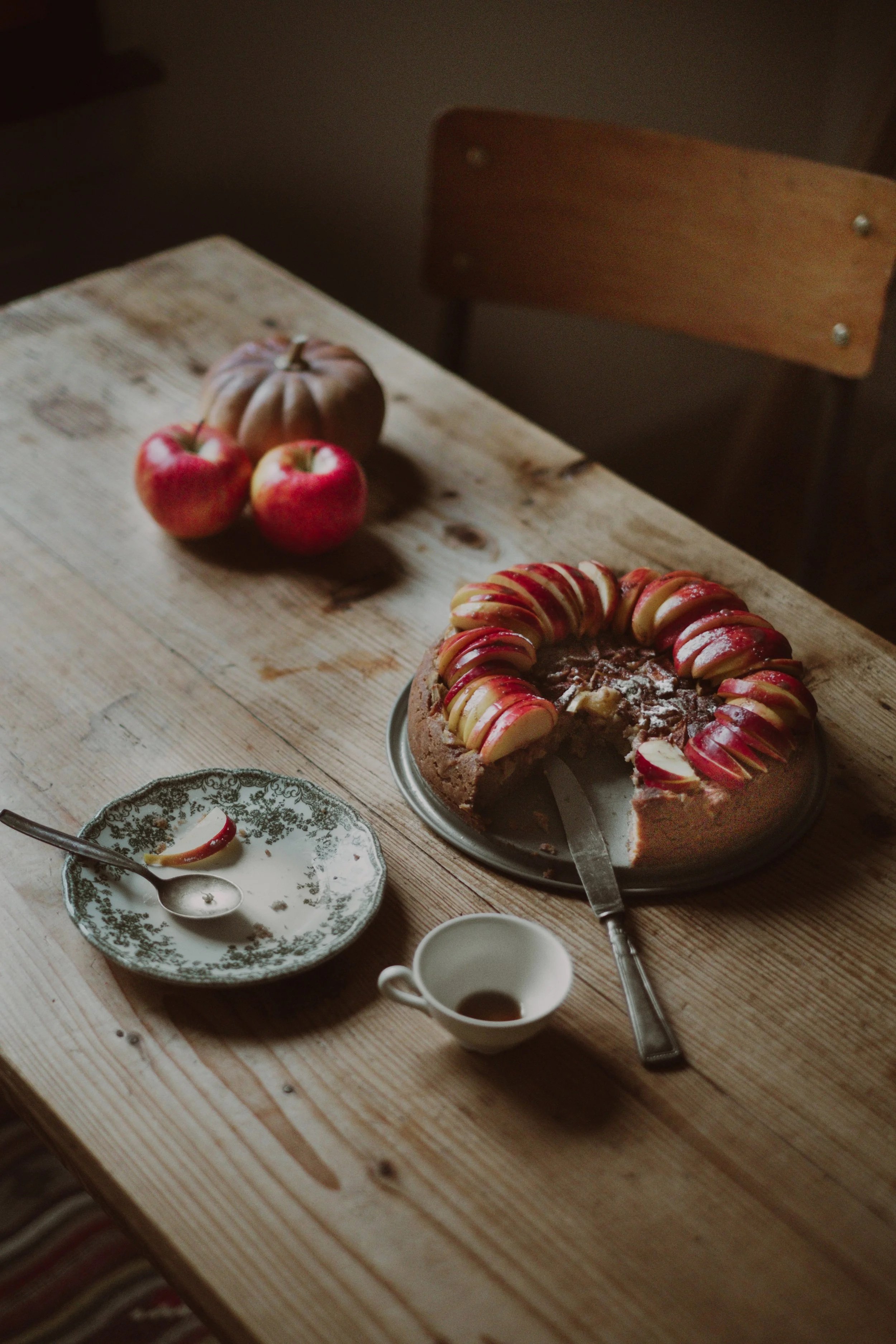 A partially eaten apple and a whole apple on a wooden table, with a homemade apple cake topped with sliced apples and red syrup, a small plate with a spoon and apple slice, a small cup, and a pumpkin in the background.