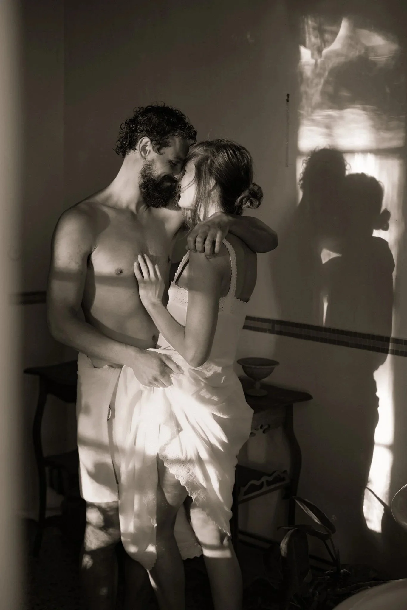A black and white photo of a shirtless man with a beard and curly hair, and a woman in a light-colored dress, with their foreheads touching and eyes closed, standing close together in a dimly lit room. Shadows of their faces are cast on the wall behi