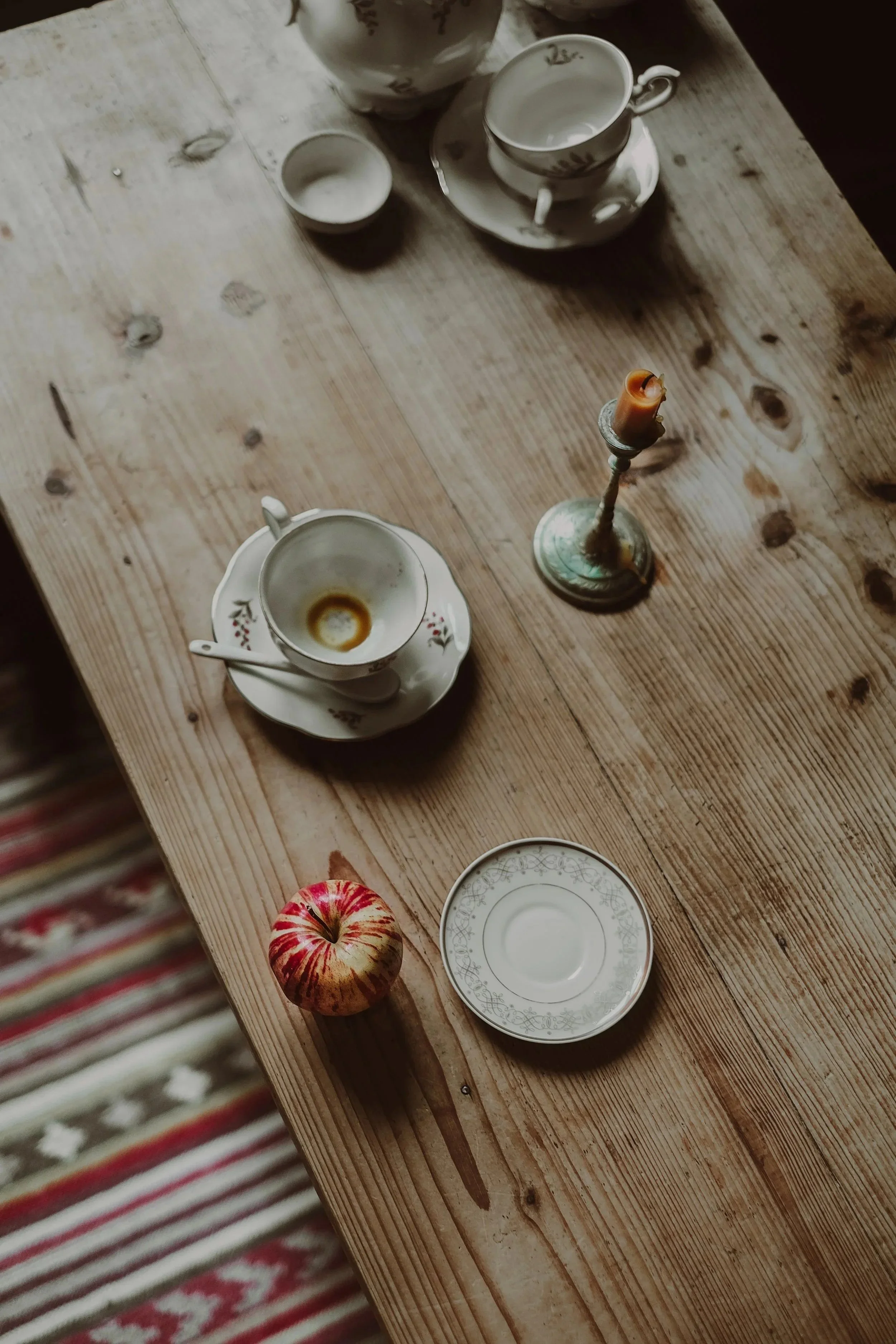 A rustic wooden table with a dessert apple, a white teacup with a floral pattern, an empty small bowl, a teacup with a saucer, and a candlestick with a partially melted candle.