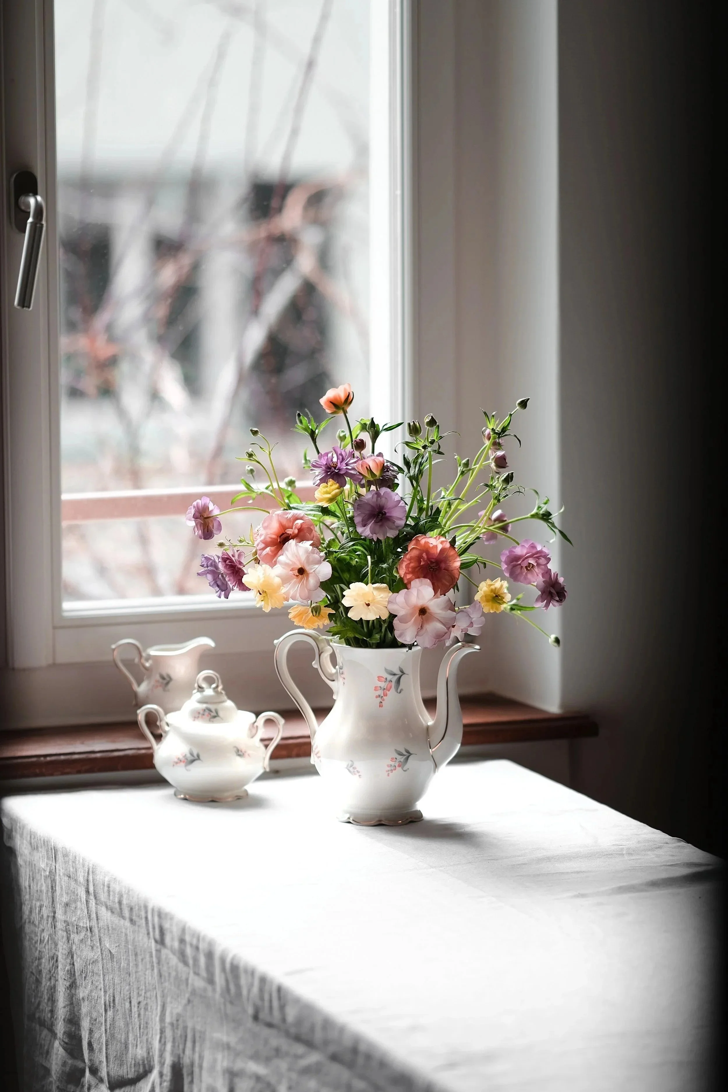 A window with a white ceramic teapot vase filled with colorful flowers, including pink, purple, and yellow blooms, on a white tablecloth-covered table indoors.