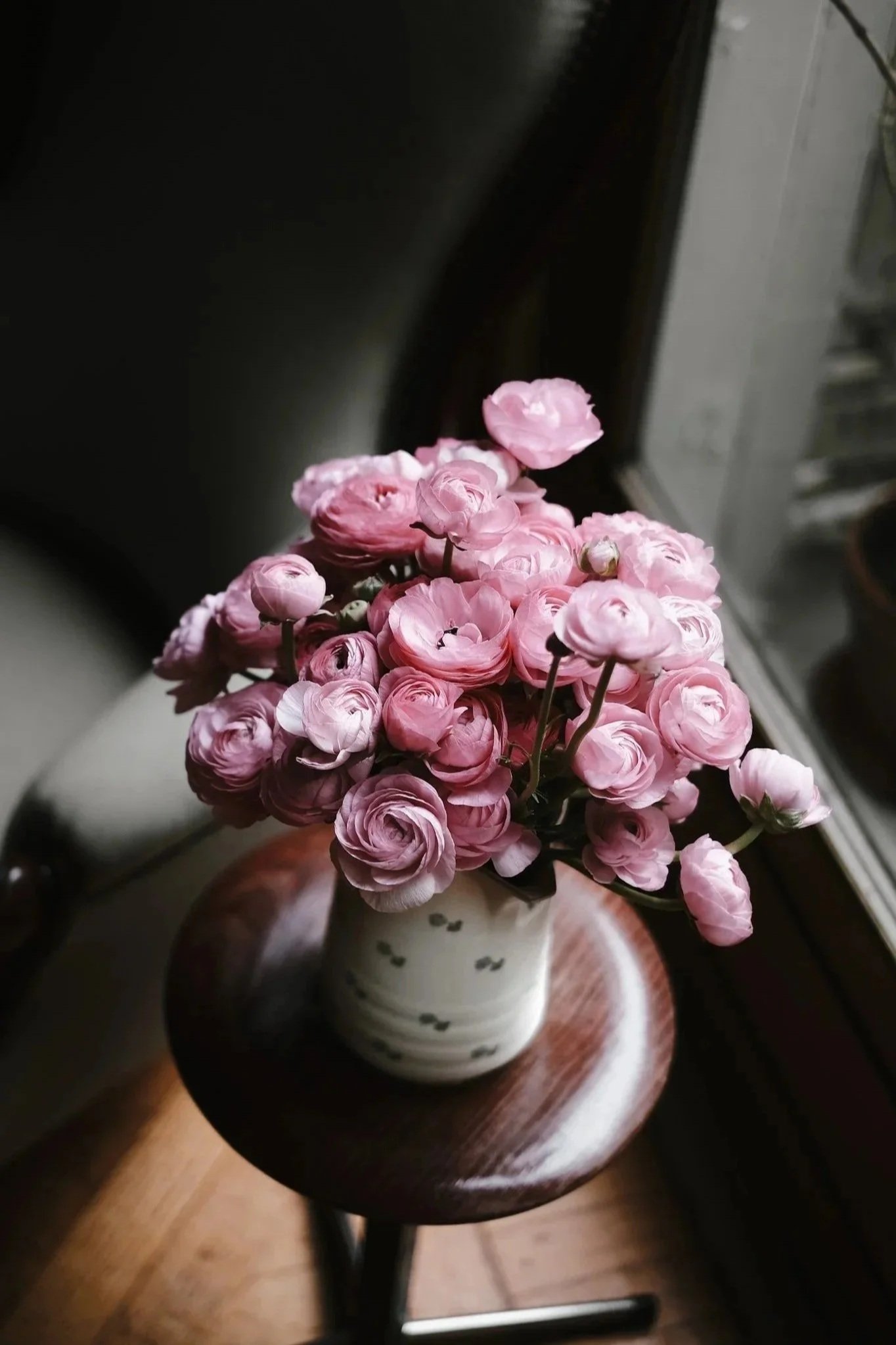 A bouquet of pink ranunculus flowers in a white ceramic vase with black spots, placed on a round wooden table next to a window.
