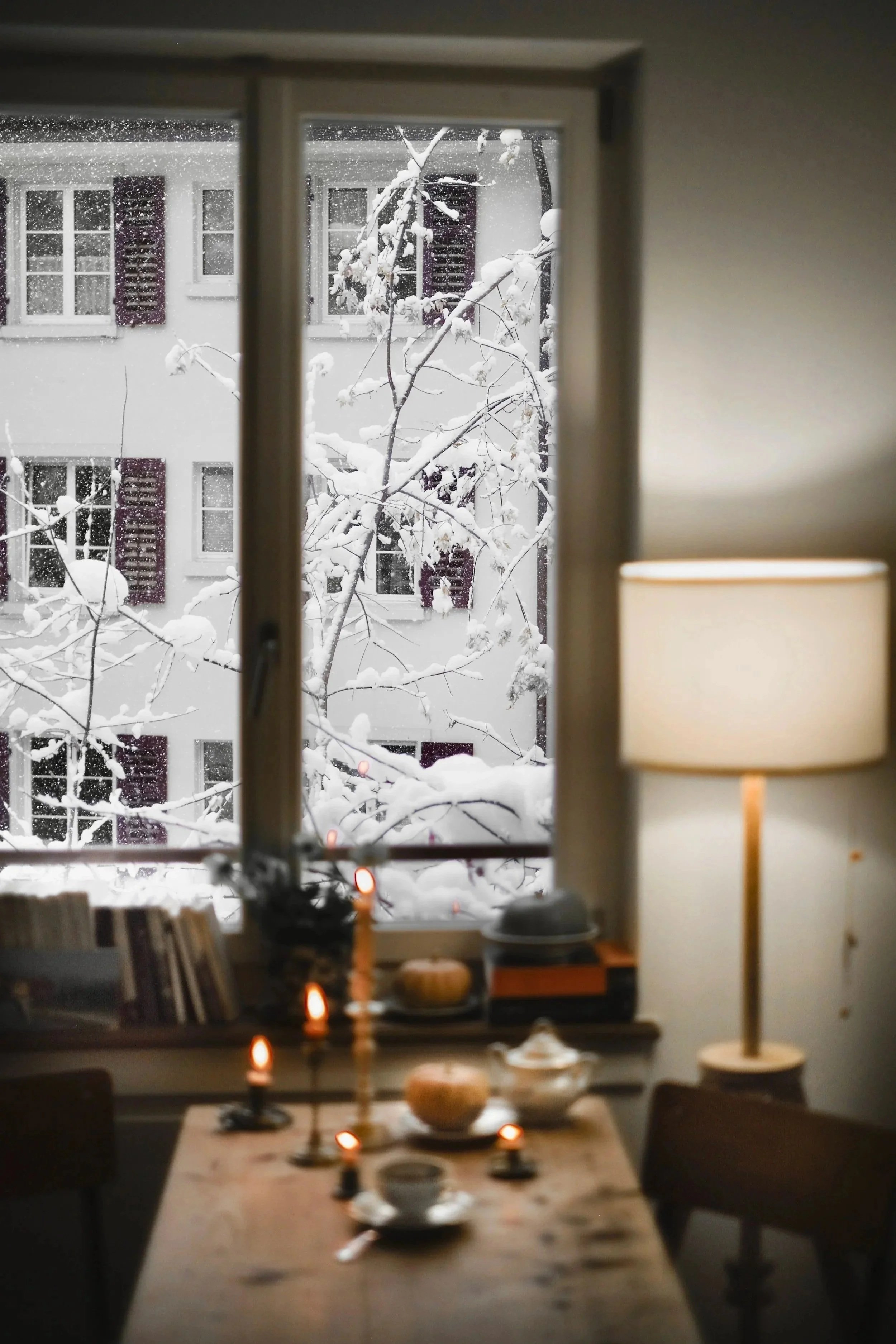 Indoor scene with a table set for dessert next to a window showing a snowy outdoor scene with snow-covered tree branches and a building with multiple windows.
