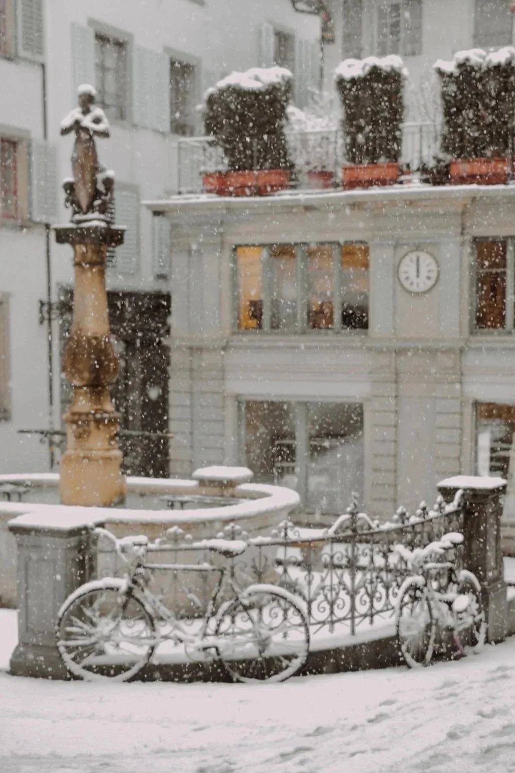 Snow falling on a fountain, a decorative bicycle, and a building with a clock and potted plants on a balcony.