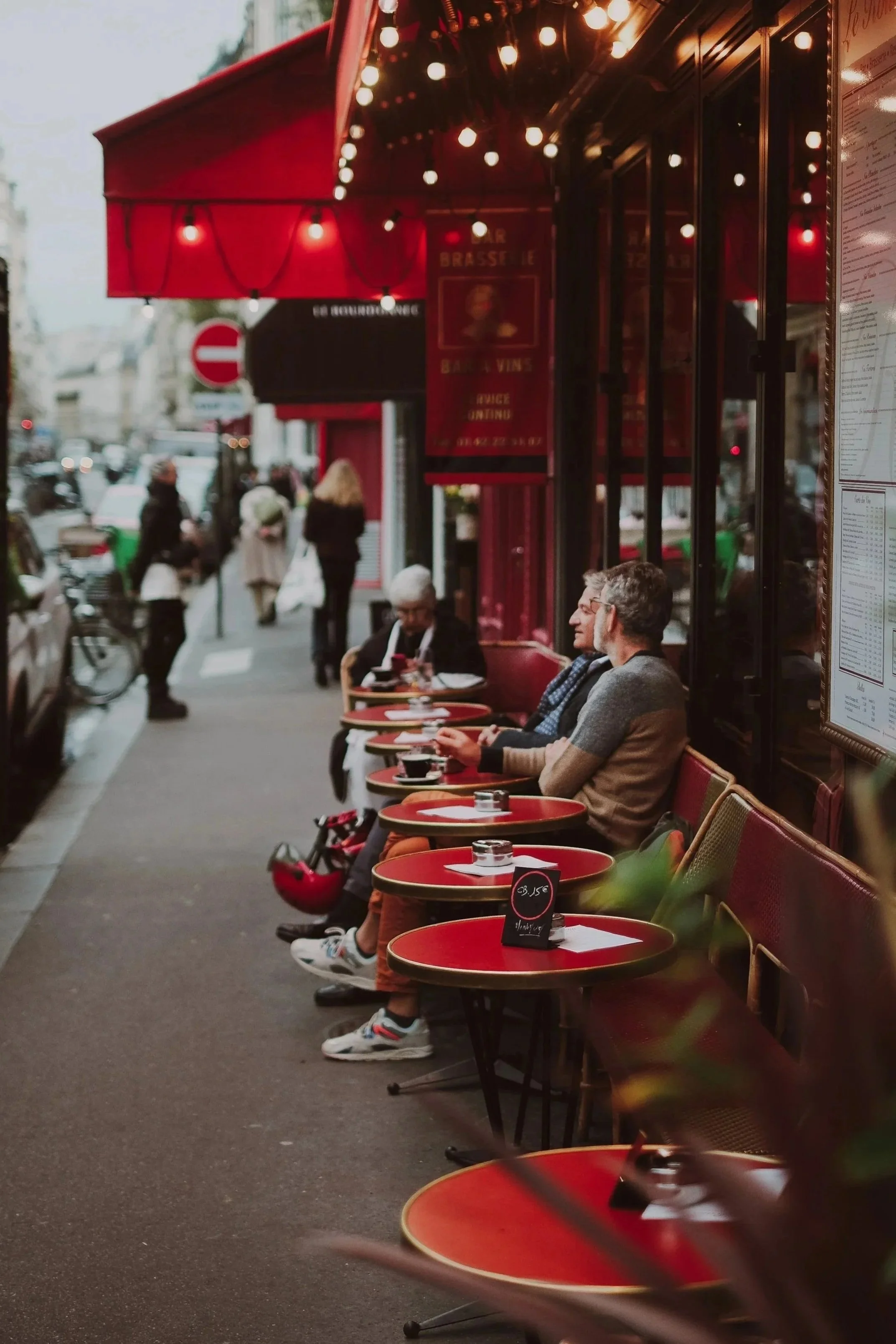 Parisian sidewalk cafe with red tables, chairs, and awning, people sitting and chatting, some walking by, illuminated with warm string lights.