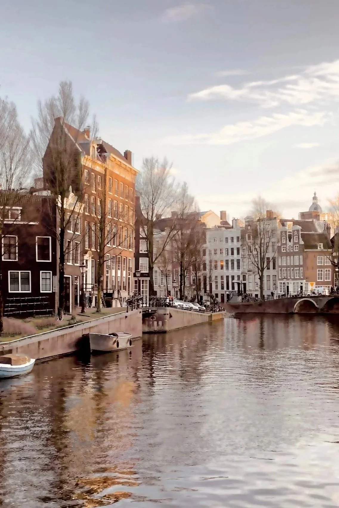 Scenic view of a canal with boats, historic buildings, and a bridge in a European city, likely Amsterdam, during sunset.