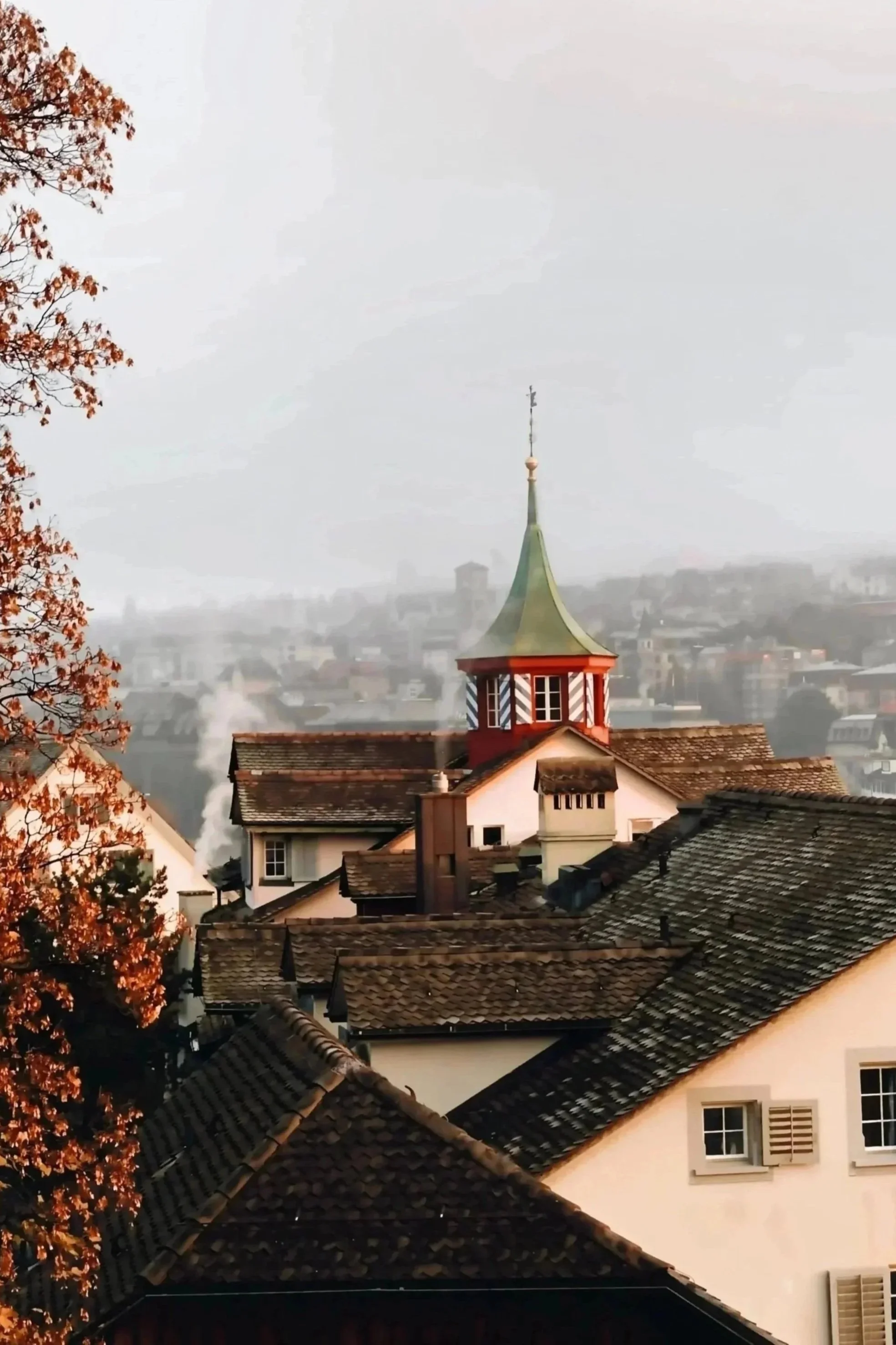 Autumn scene of rooftops and a church steeple in a foggy city with a tree with orange leaves in foreground.