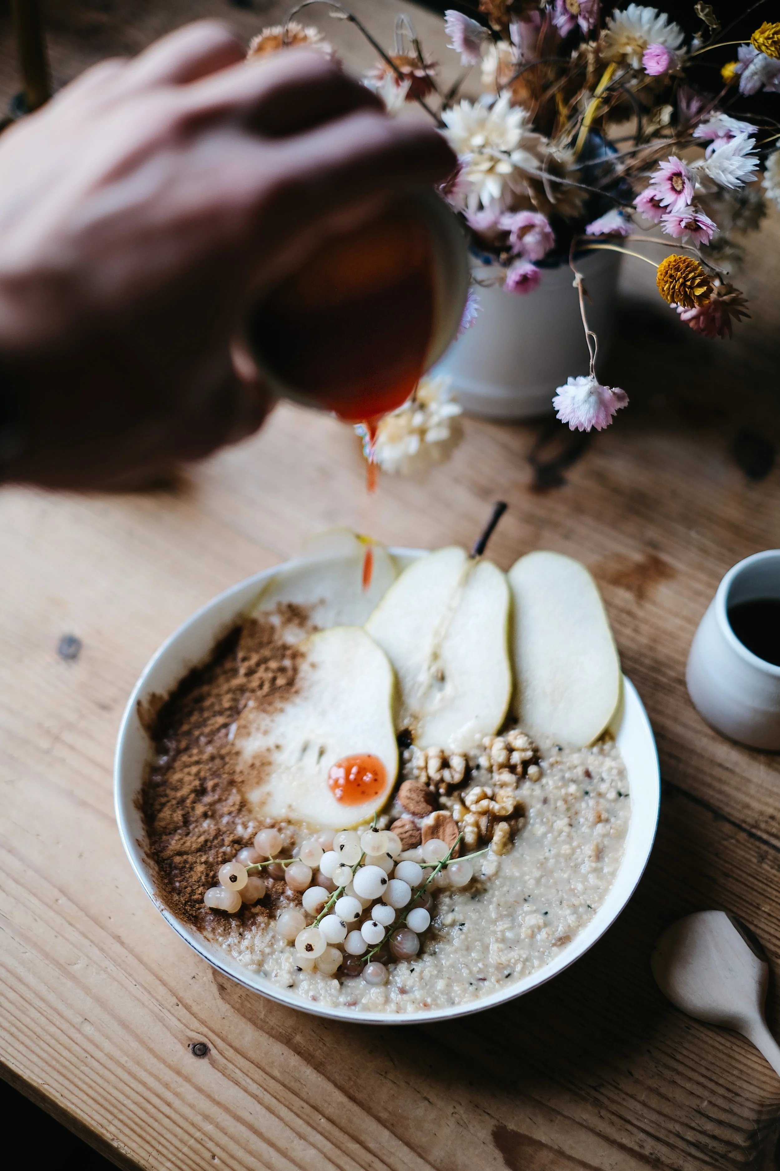 A bowl of oatmeal topped with sliced pears, a poached egg, white currants, chopped nuts, and brown sugar on a wooden table. A person is pouring syrup over the oatmeal, a cup of coffee is nearby, and a vase of pink and white flowers is in the backgrou