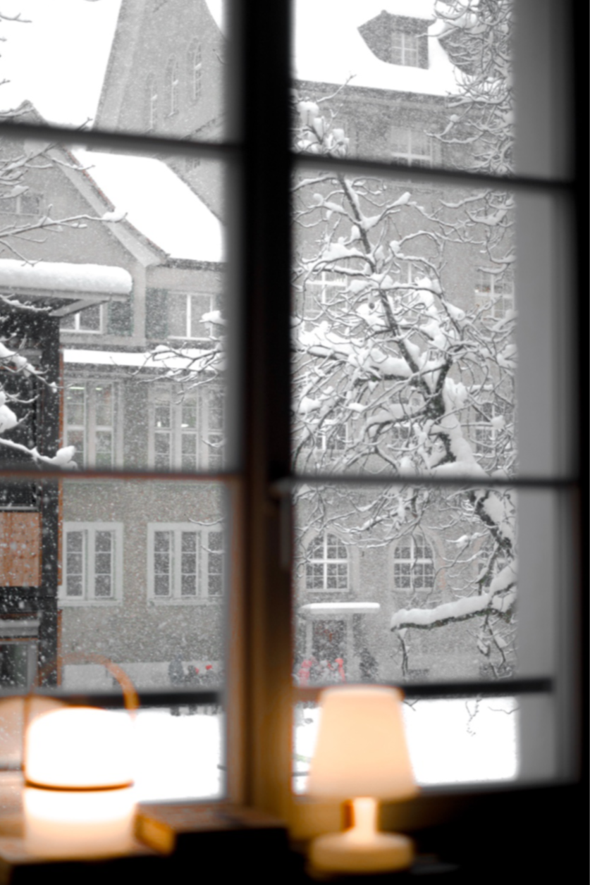 View from inside a house looking out a window at a snowy scene with snow-covered trees and buildings outside. Two small lamps are turned on on the windowsill.