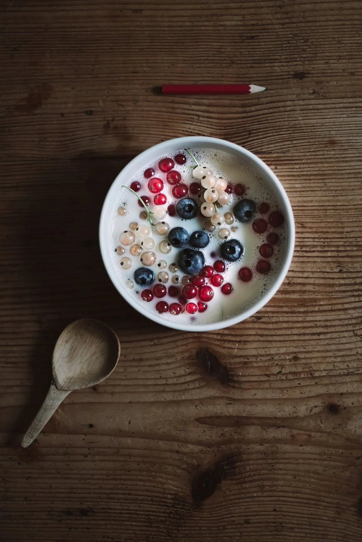 A bowl of milk topped with blueberries, red currants, and white currants on a wooden table, with a wooden spoon and a red pencil nearby.