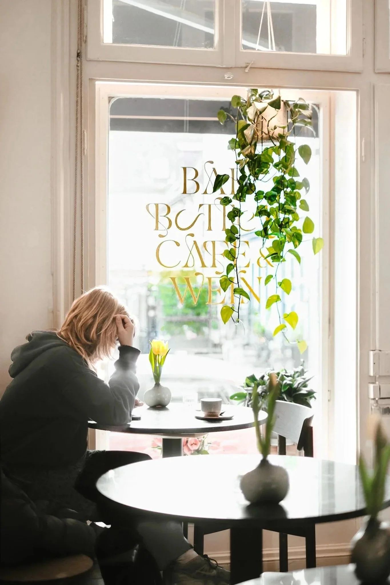 A woman sitting alone at a small table in a cafe near a window with hanging plants and a vase with yellow tulips, holding her head in her hand.