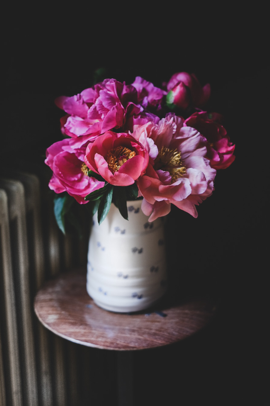 A bouquet of pink peonies in a white vase with blue accents on a wooden side table, against a dark background.