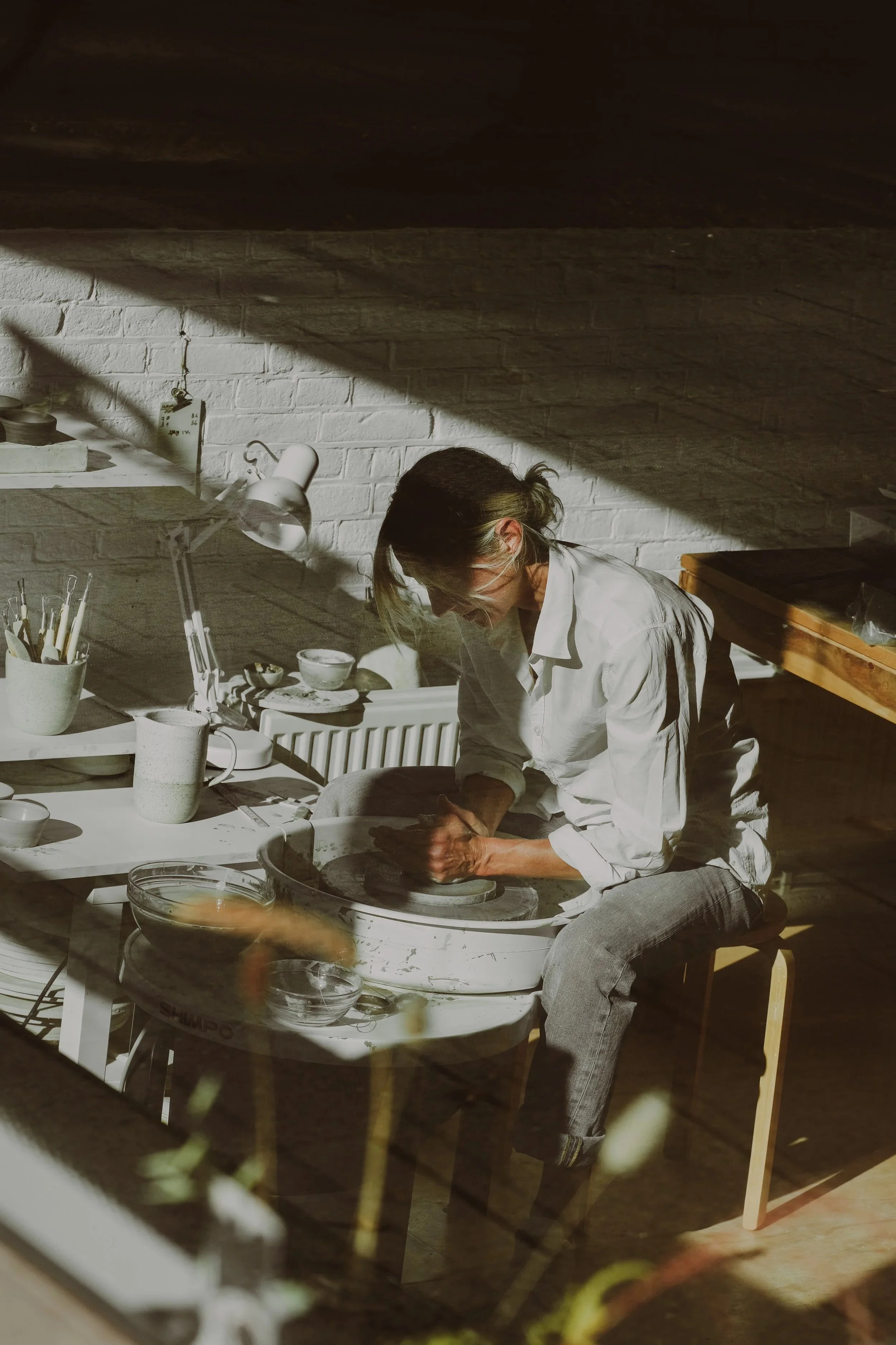 A woman sitting on a stool in a light-filled room, working with pottery on a pottery wheel surrounded by pottery tools, cups, bowls, and a lamp.