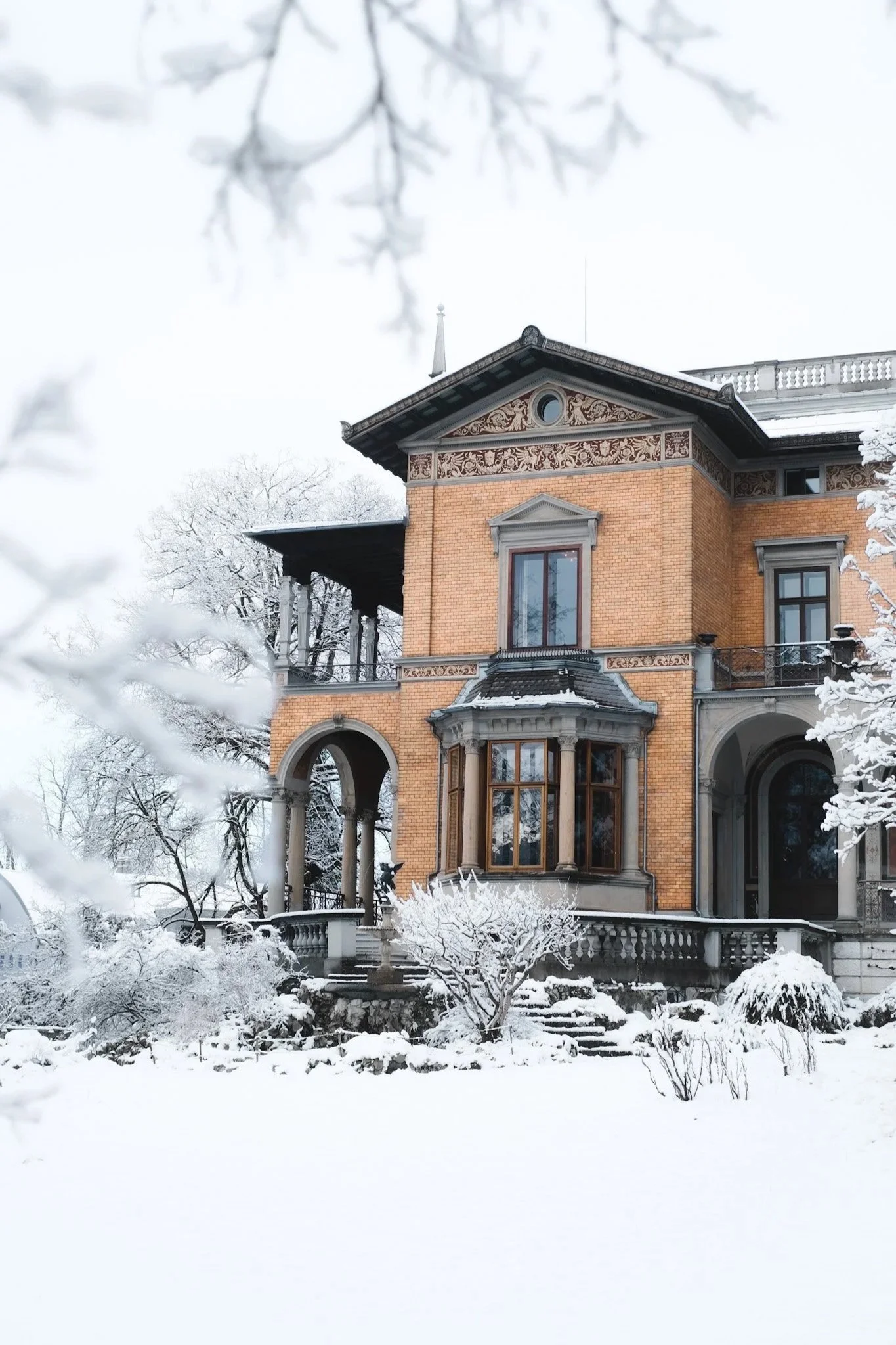 A large historic house with brick walls, ornate architectural details, and tall windows, surrounded by snow-covered trees and ground in winter.